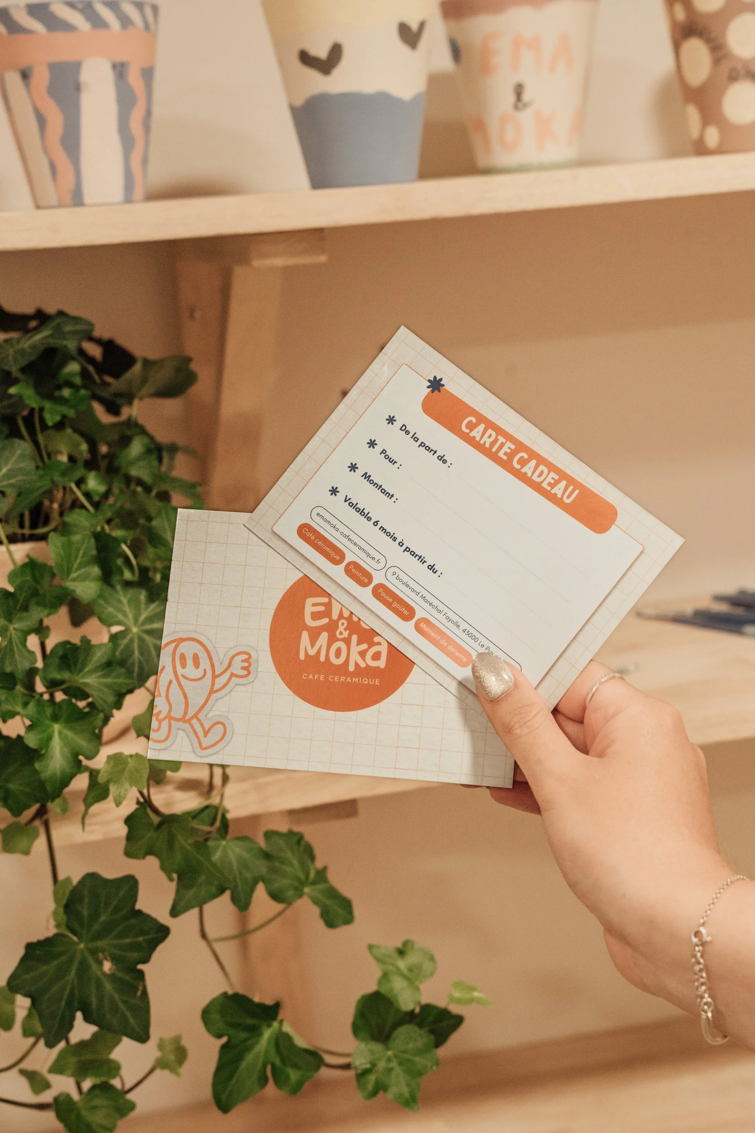 Une main tient des cartes-cadeaux de la boutique Ew&Moka, avec des pots décorés en arrière-plan.