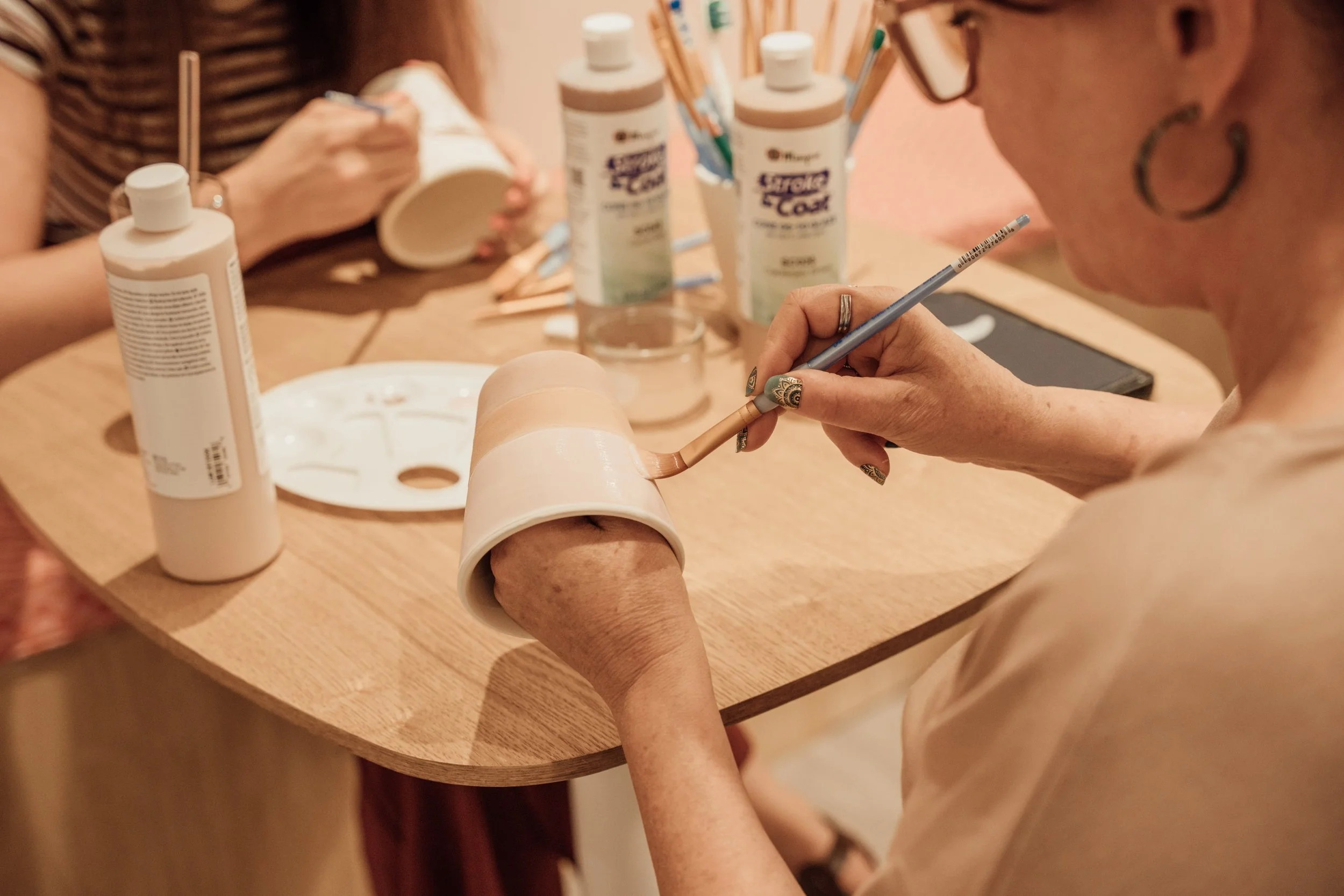 Une personne peignant un objet en utilisant un pinceau doté d'une peinture beige ou chair. Sur la table en bois, il y a plusieurs bouteilles de colle et d'autres fournitures, ainsi qu'une main tenant un vide-ordures.