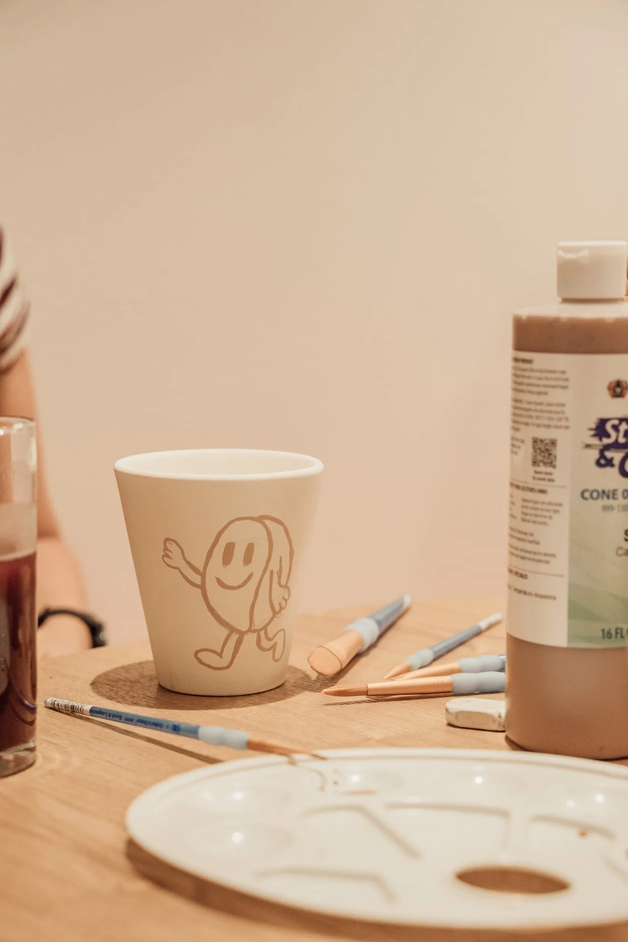 Une tasse en papier avec un dessin d'une character cheerful, entourée d'outils d'art tels que des pinceaux, des stylos et un tube de peinture, sur une table en bois dans un environnement créatif.
