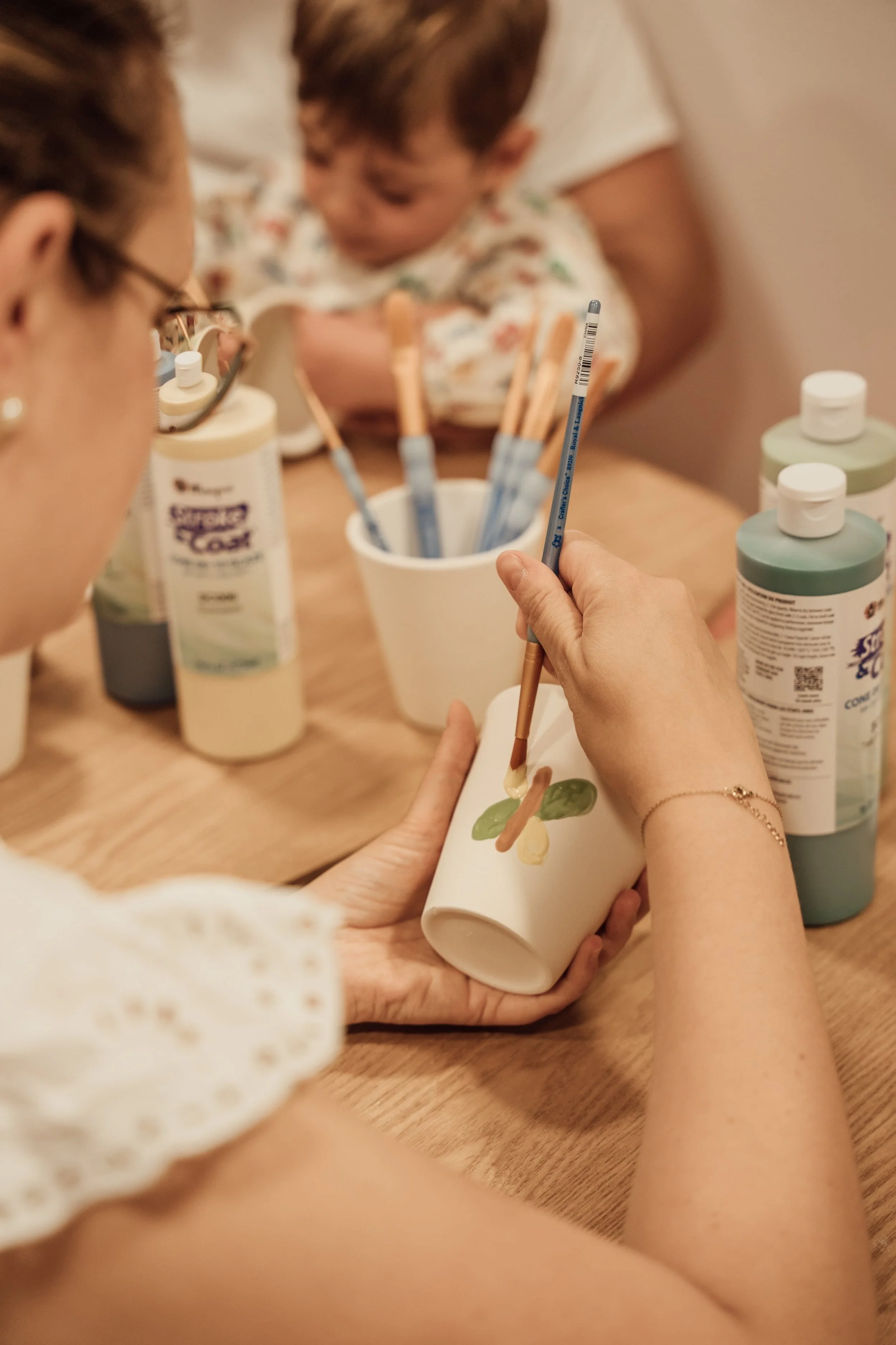 Une personne peint un vase blanc avec des motifs colorés, entourée de plusieurs bouteilles de peinture et de pinceaux sur une table en bois.