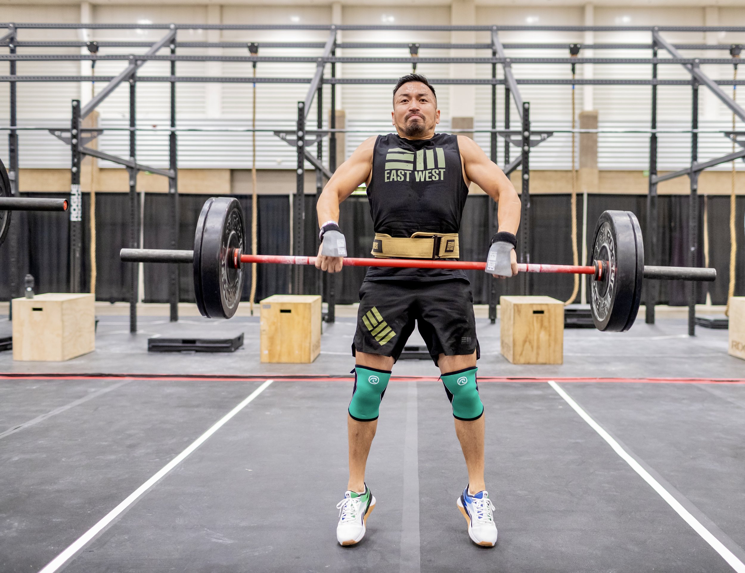 A man in athletic wear lifting a barbell in a gym with black flooring and gym equipment in the background.