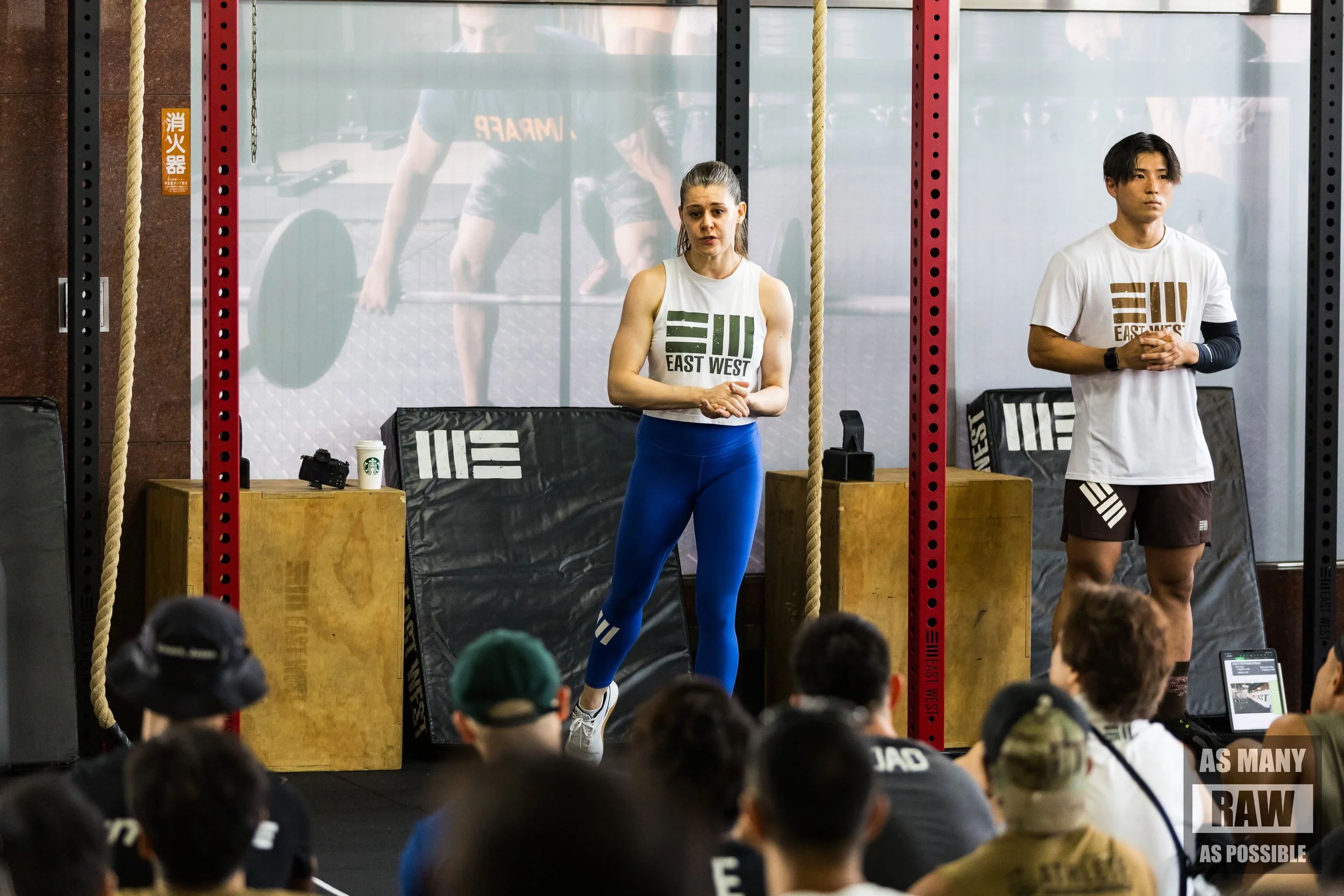 A woman and a man standing in front of a crowd at a gym, wearing matching 'East West' t-shirts. The woman is speaking and gesturing with her hands, while the man stands with his hands clasped. There are gym equipment and a large poster with a person lifting weights in the background.