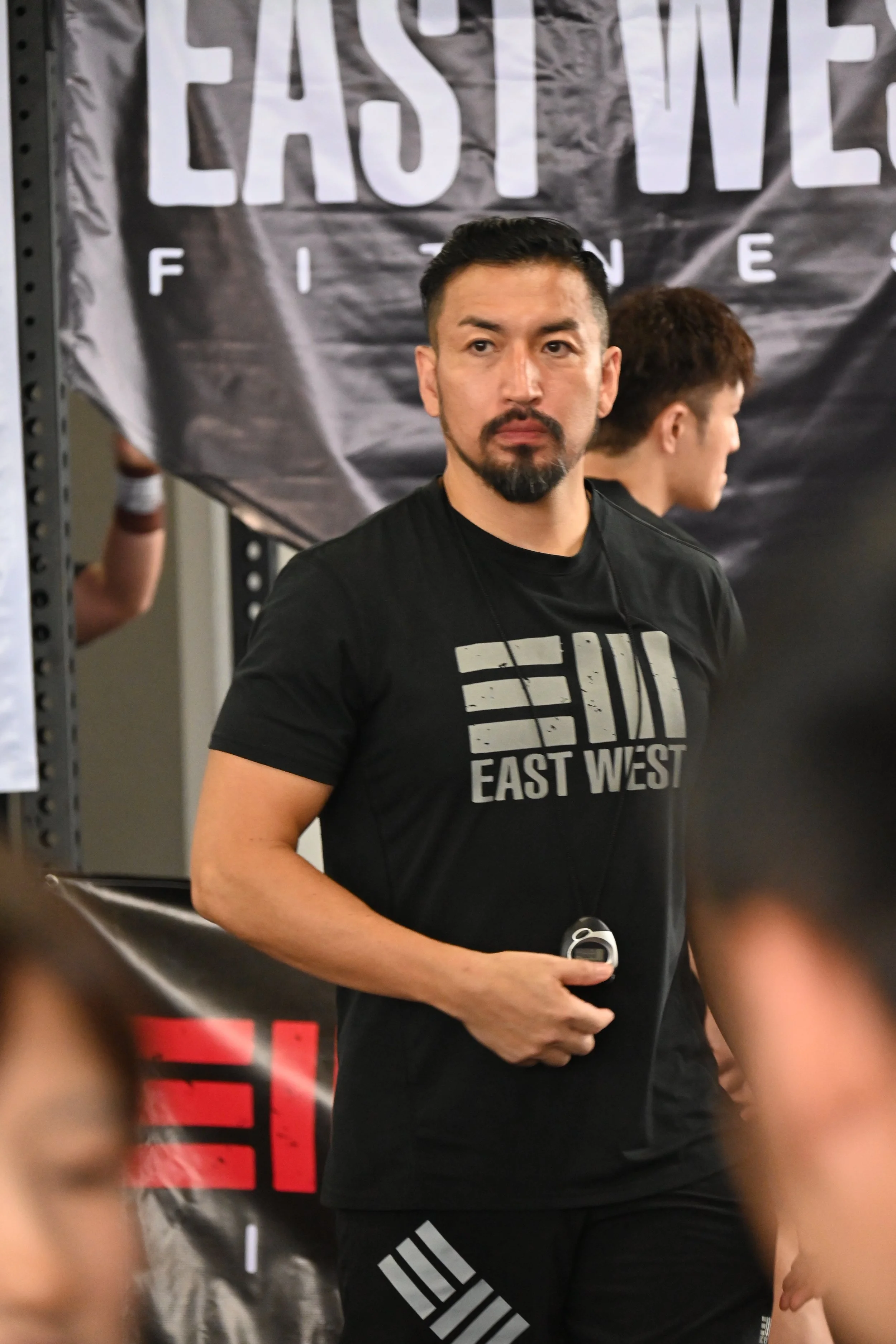 A man with short black hair, a beard, and mustache wearing a black T-shirt with the words 'EAST WEST' and riding a black Adidas shorts is holding an object in his right hand. Behind him is a black banner with the words 'EAST VILL' visible. There are other people partially visible in the background.