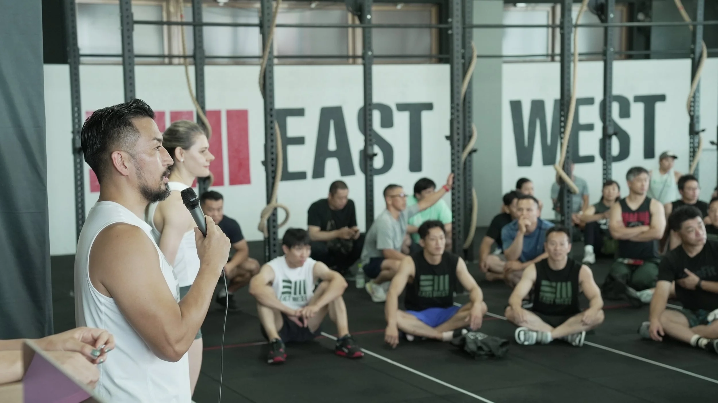 People gathered in a gymnasium, sitting on the floor, listening to a man with a microphone, with a large sign in the background that reads 'EAST' and 'WEST'.