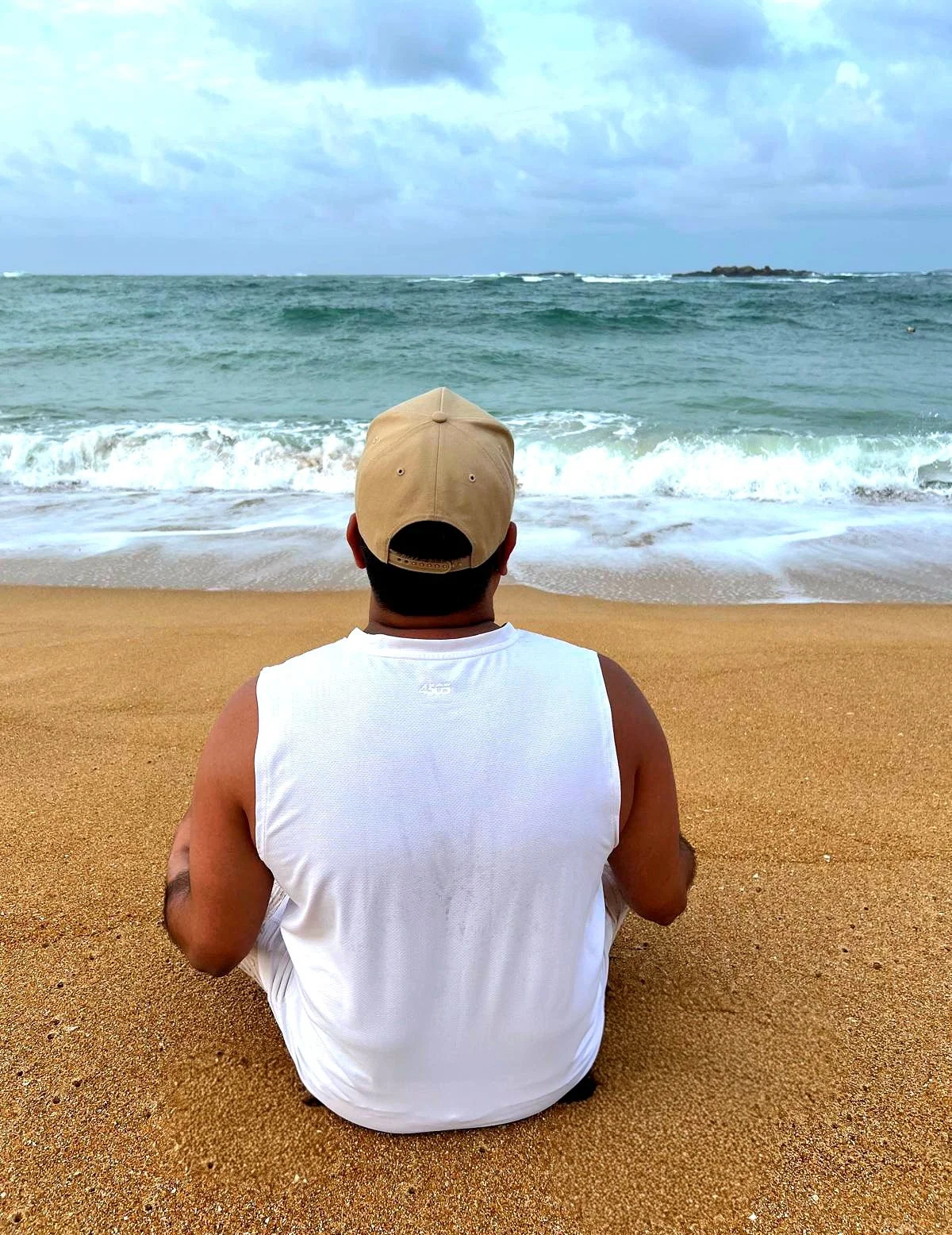 Sankha Senanayake sitting on the sandy beach facing the ocean, wearing a beige cap and a white tank top, with waves approaching the shoreline under a cloudy sky.