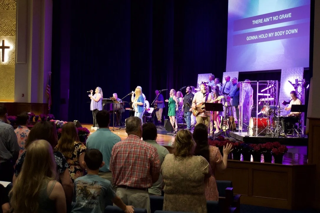 Fairmount Church congregation attending a worship service with a band and singers performing on stage, lyrics displayed on a large screen.