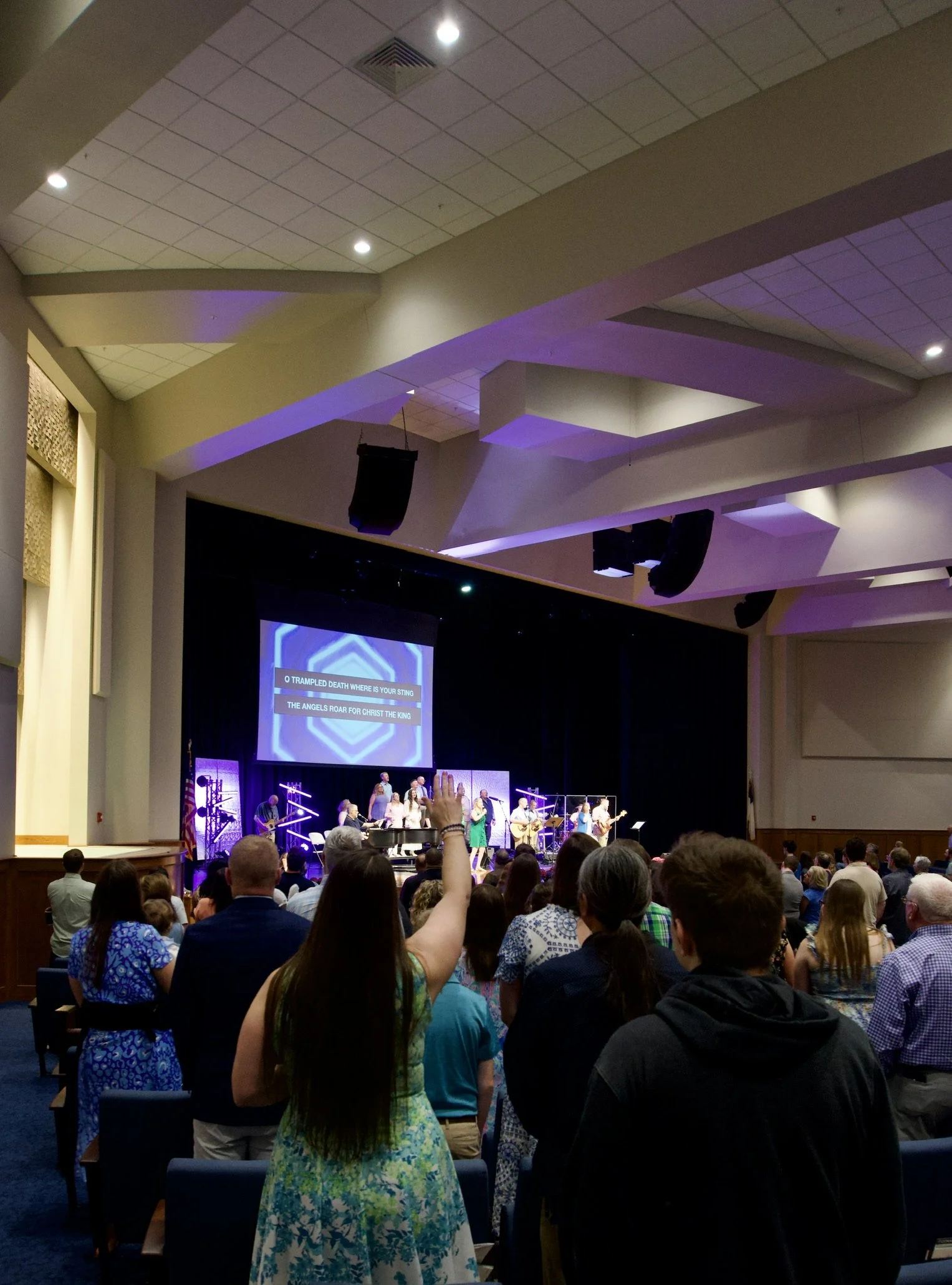 Audience in a church or auditorium watching a worship band and choir perform on stage, with some people raising their hands, and a large screen displaying song lyrics.