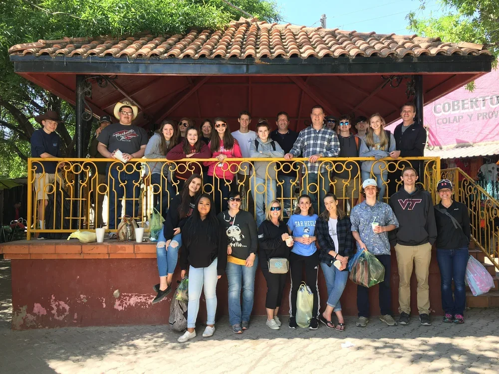 Group of people gathered on a decorated outdoor gazebo with a terracotta tiled roof, some standing on the upper level and others on the ground in front, smiling for the photo, with trees and a pink-colored wall in the background.