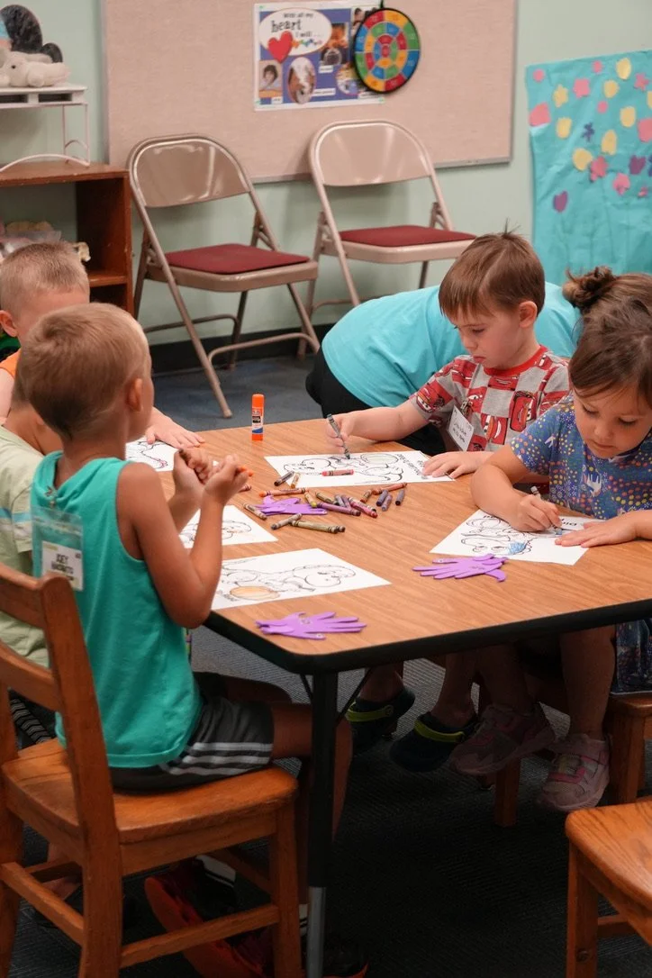 Children sitting at a table coloring paper with crayons, in a classroom setting.
