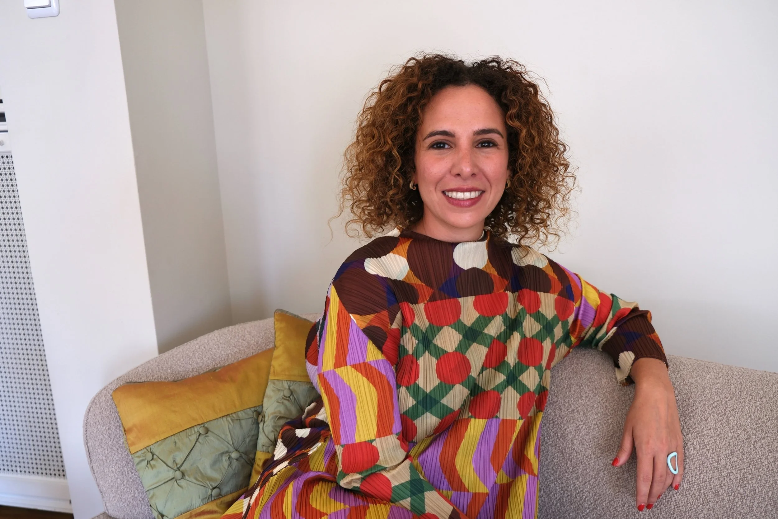 A woman with curly hair smiling while sitting on a beige sofa in a room with white walls. She is wearing a colorful dress with a geometric pattern. There are yellow and light green pillows on the sofa.