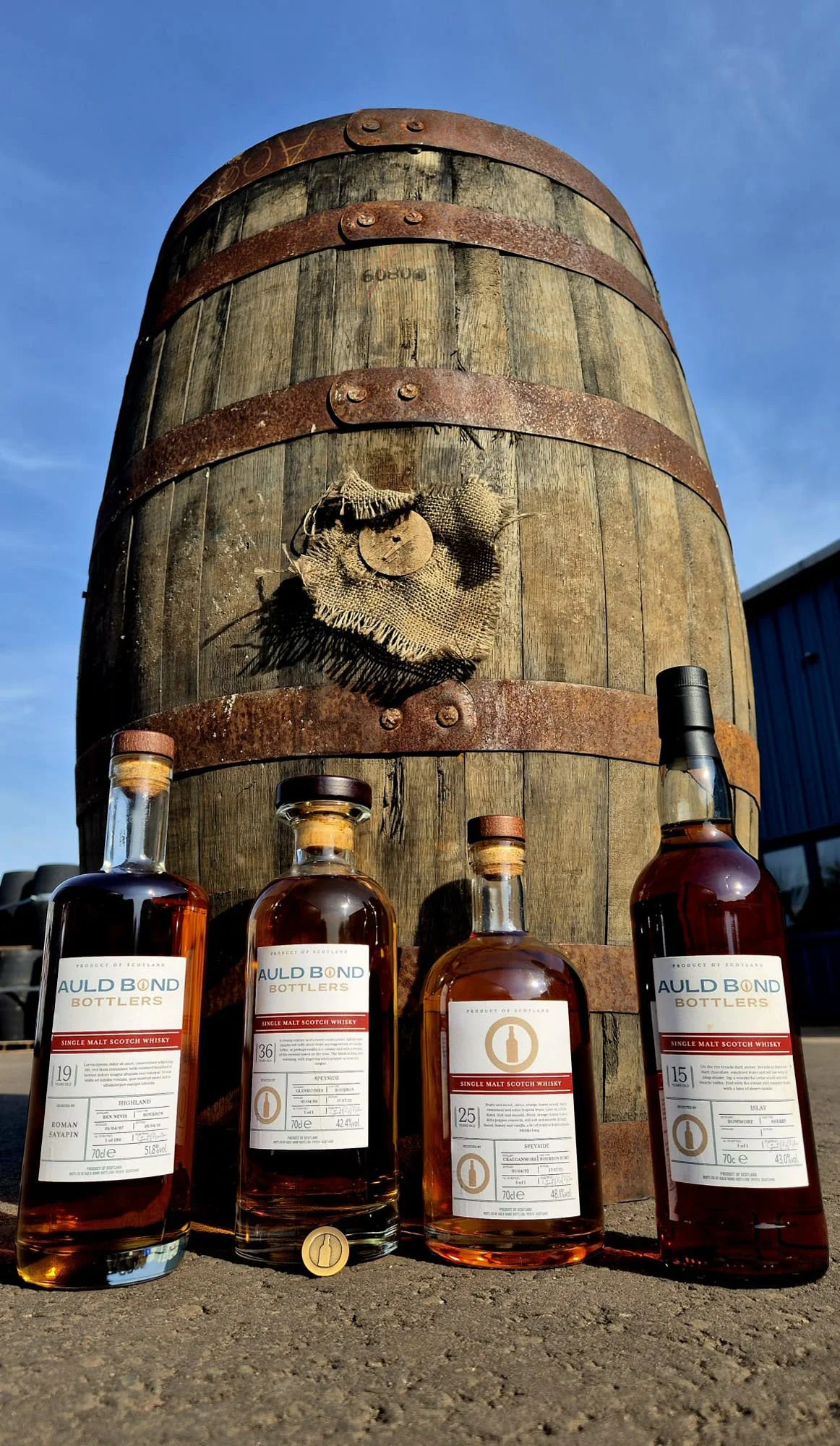 Four bottles of single malt Scotch whisky branded Auld Bond Bottlers are placed on the ground in front of a large, weathered wooden barrel with rusted metal bands. The barrel has a piece of burlap cloth attached to it, and the background shows a clear blue sky and part of a building.