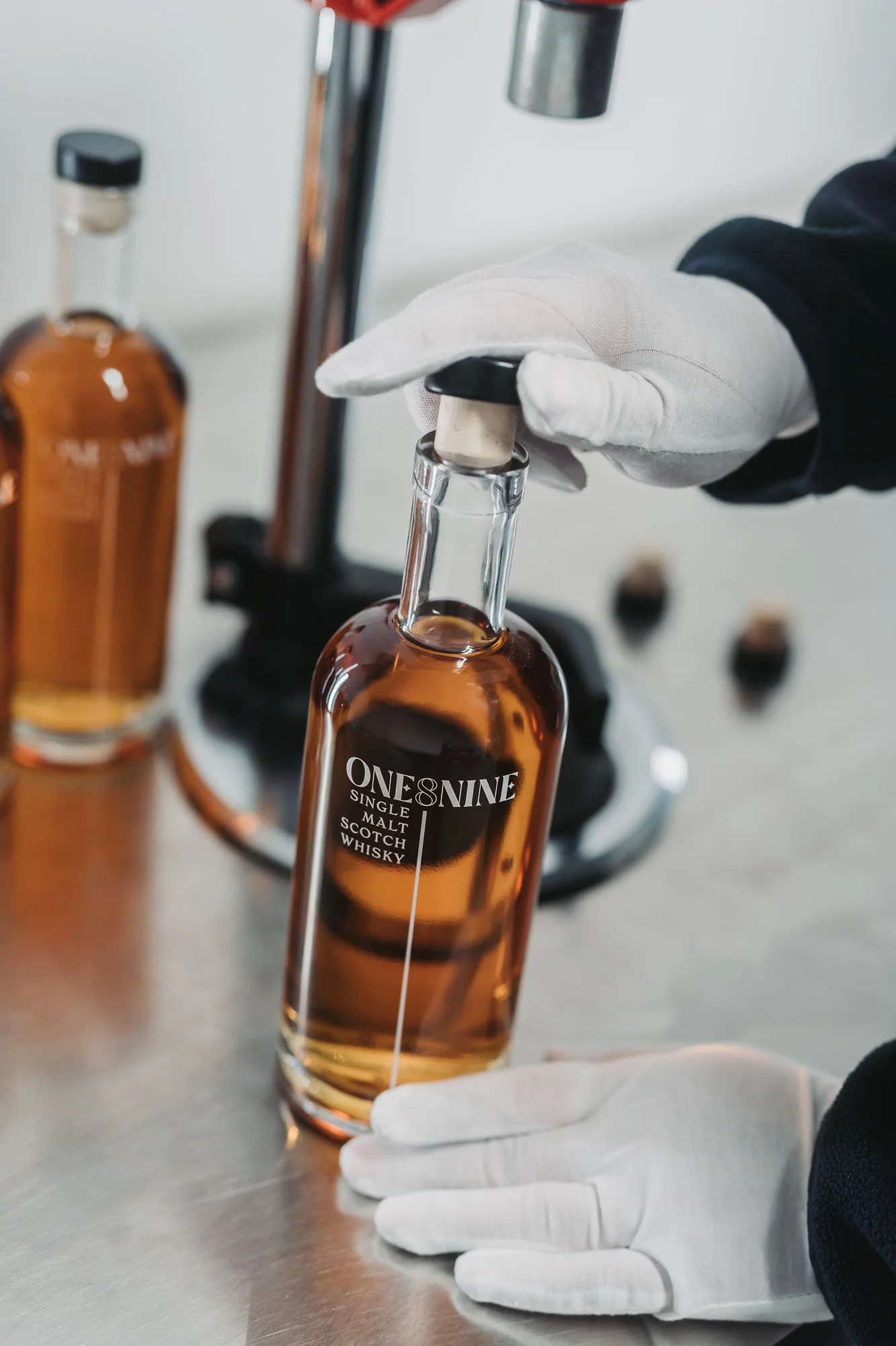 A person wearing white gloves handling a bottle of whiskey in a laboratory or distillery setting. The bottle is labeled 'ONE & NINE SINGLE MALT SCOTCH WHISKY' and is being examined or filled.