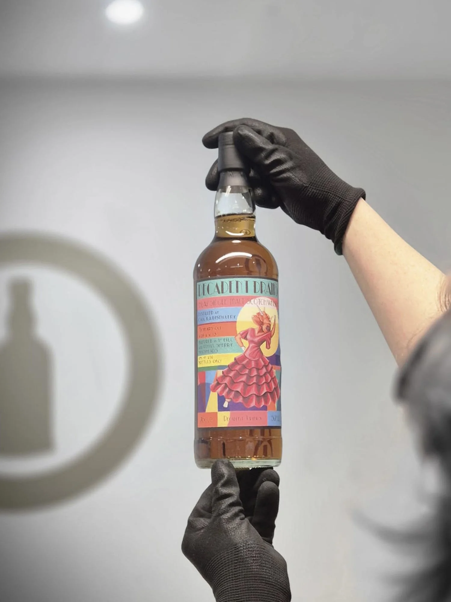A hand wearing a black glove holding a glass bottle with a colorful label featuring an illustration of a woman in a pink dress. The background is plain and light-colored.