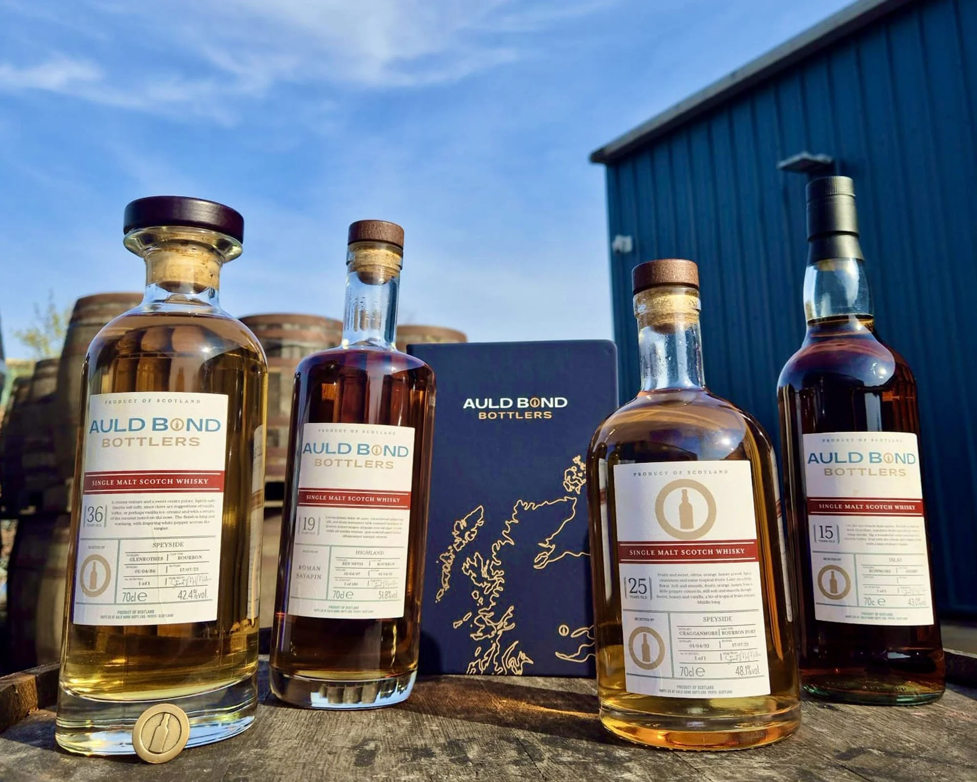 Several bottles of single malt Scotch whisky displayed outdoors with a blue building and sky in the background. The bottles are from Auld Bond Bottlers and vary in label details.