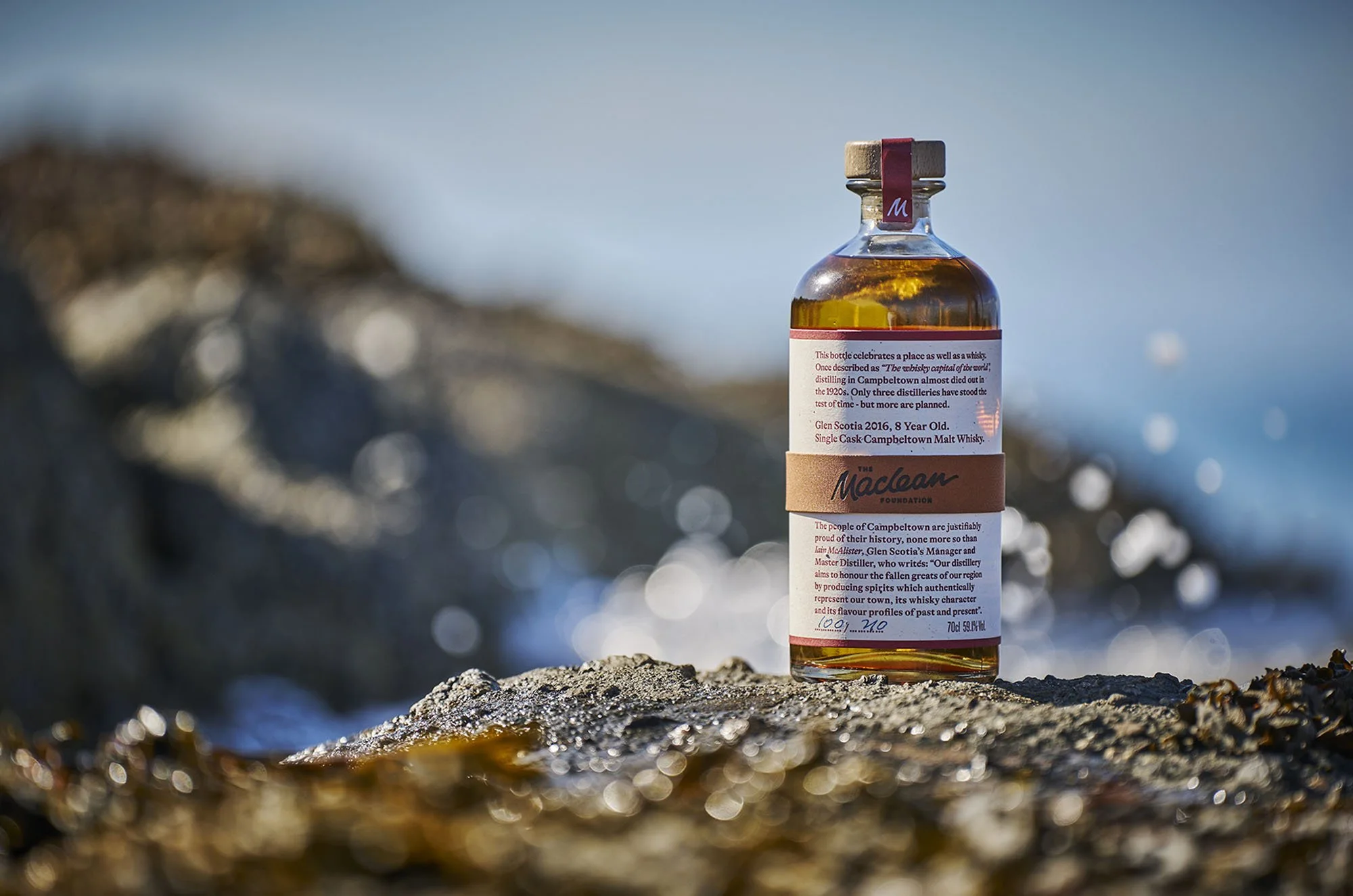 A bottle of whisky is placed on rocks with a blurred background and reflections of the sky on the surface of the whisky.