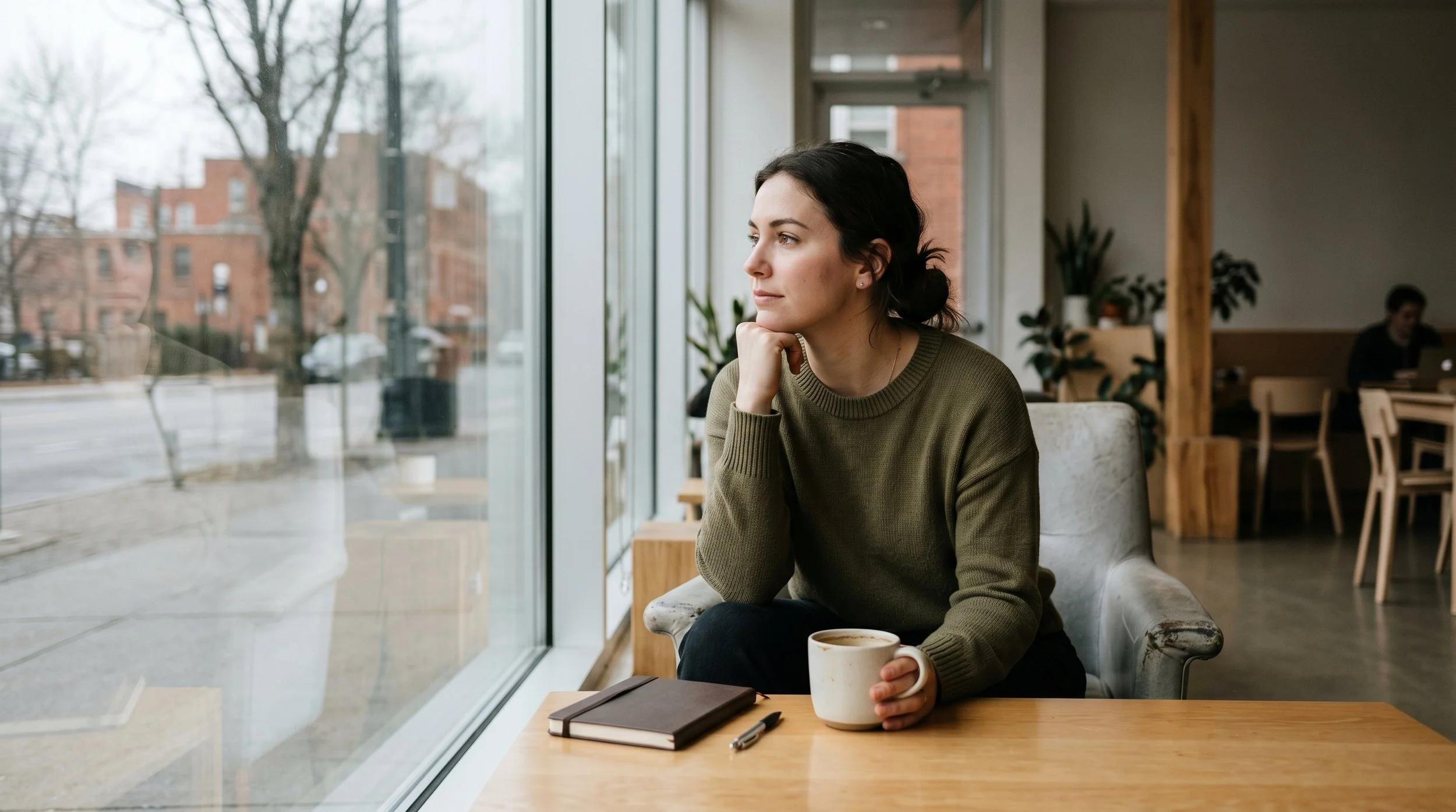 A woman with dark hair and fair skin sitting by a large window in a coffee shop, holding a cup of coffee and looking out thoughtfully. There are notebooks and a pen on the table in front of her, with a background of indoor plants and other patrons.