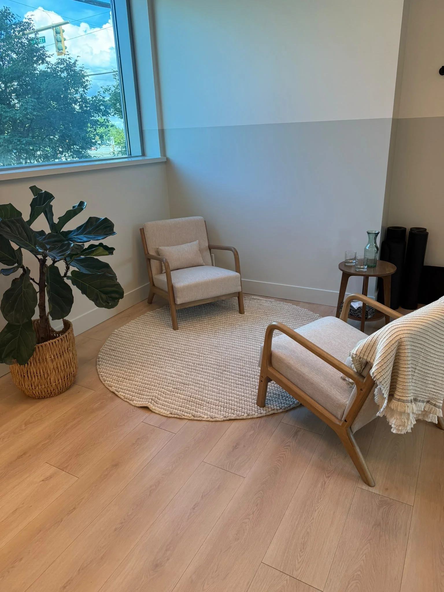 A cozy living room corner with a large window, a beige armchair, a round wooden side table with a glass carafe and glasses, a potted plant, and a woven rug on light wood flooring.