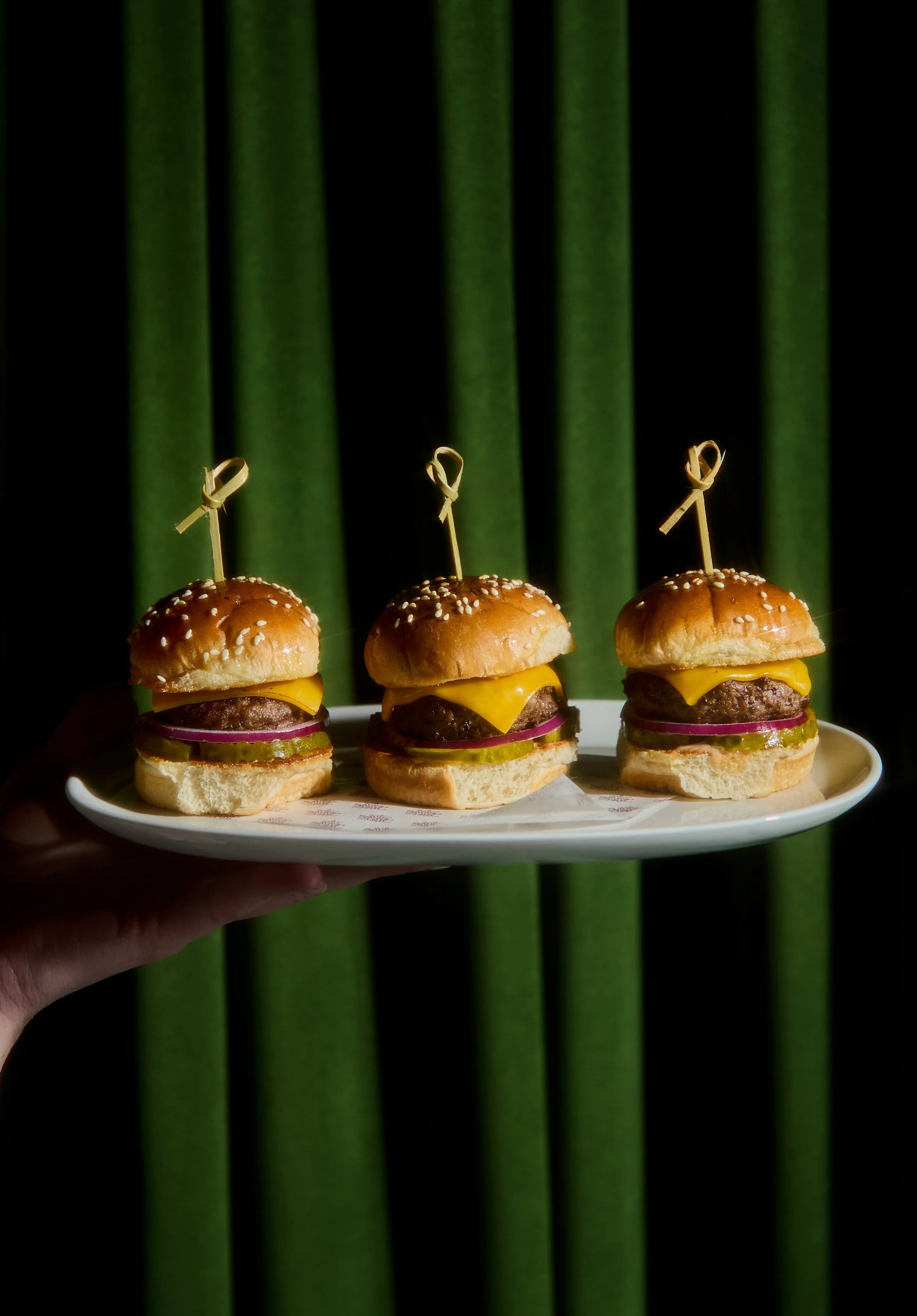 Three cheeseburgers with buns, cheese, pickles, onions, and beef patties on a white plate, with a green curtain background.