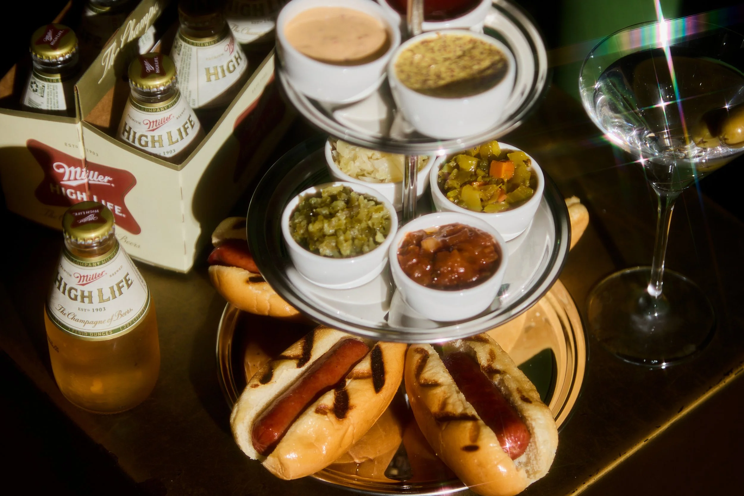 A tiered tray with small cups of chutney and sauces, cups of coffee, mini sandwiches, grilled hot dogs, and a glass of water with green olives, set on a table with Miller High Life beer bottles and packaging around.