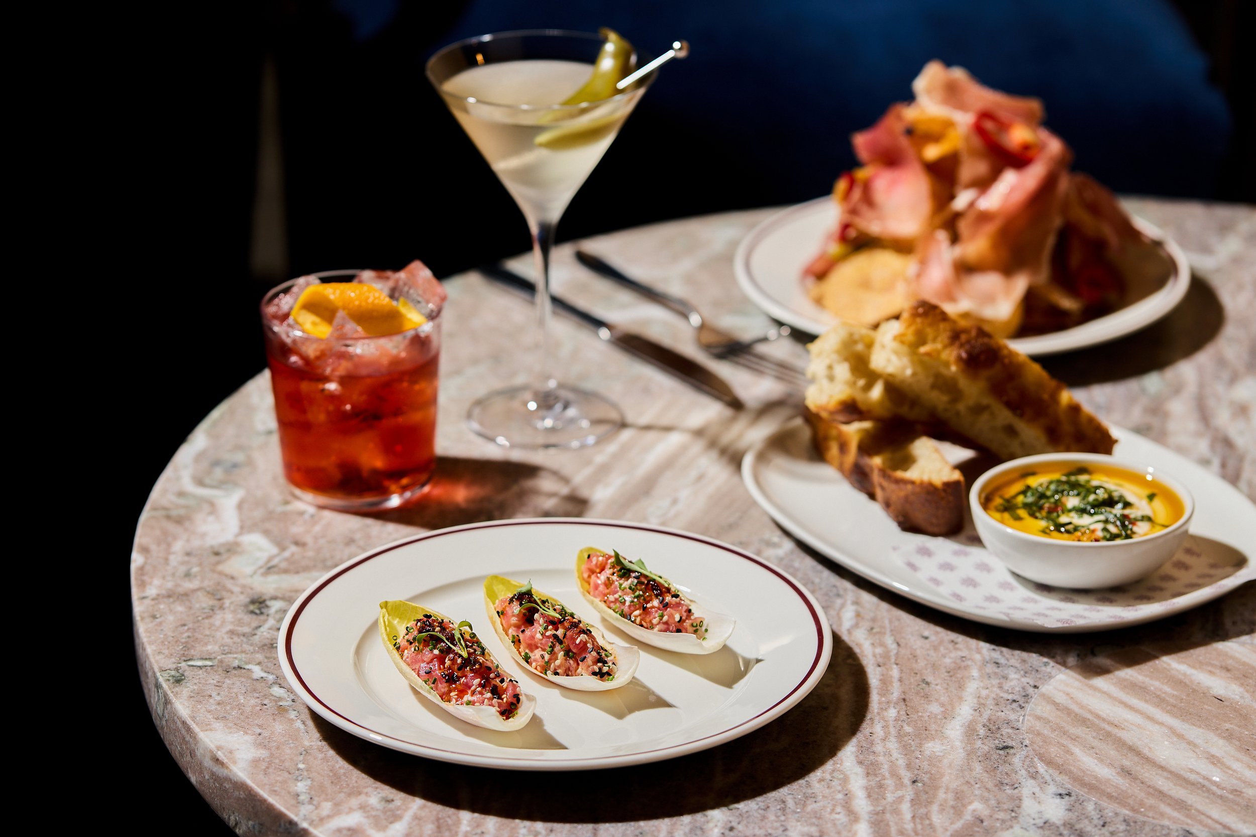 A table set with various foods and drinks, including a plate of avocado topped with fish and herbs, a small bowl of soup, a plate of sliced bread, a plate with roasted chicken and vegetables, a martini with a pickle, and a glass of red-colored cockta