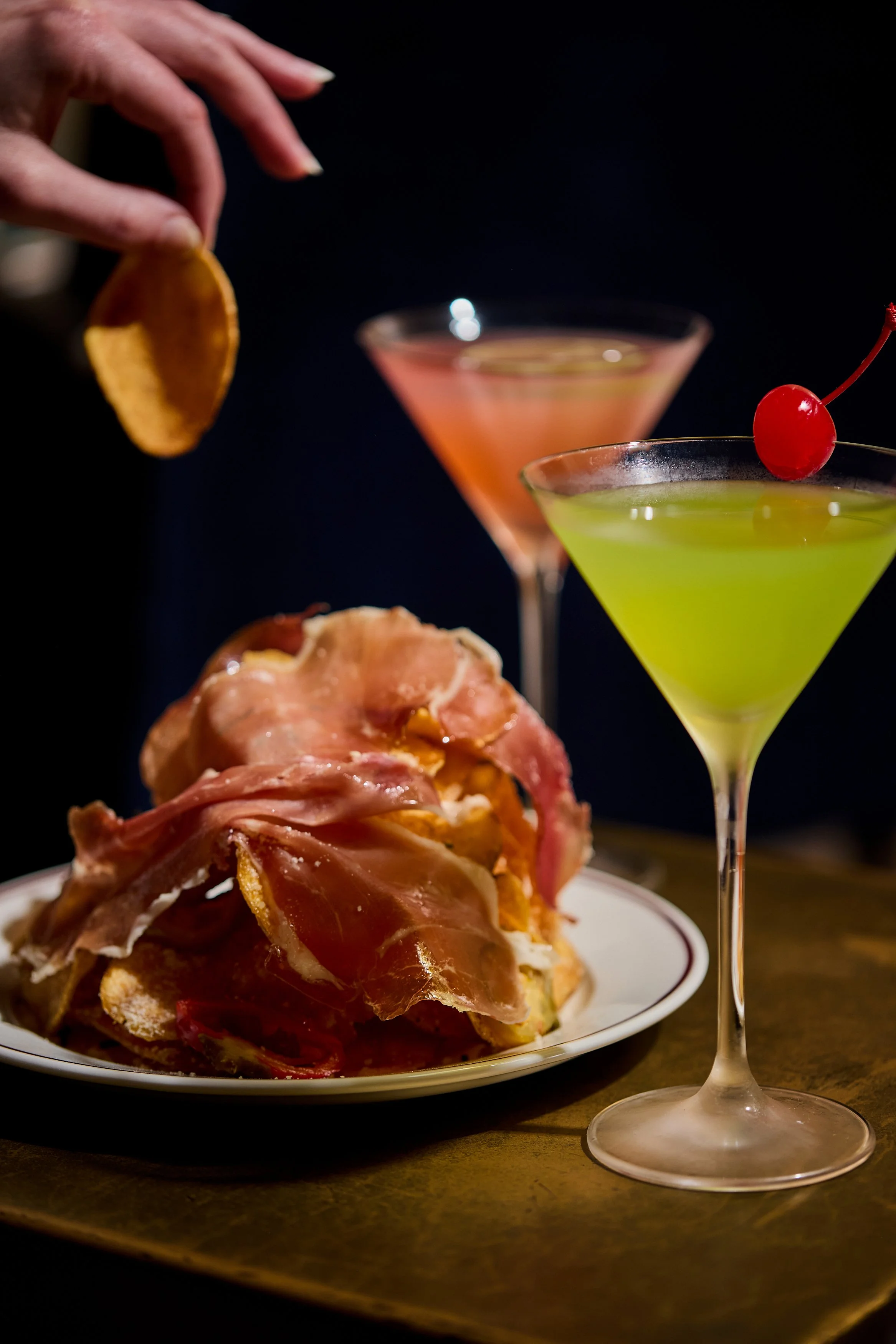 A close-up of a plate with a savory food dish topped with prosciutto, with two cocktail glasses filled with brightly colored drinks in the background, one garnished with a cherry, against a dark background.