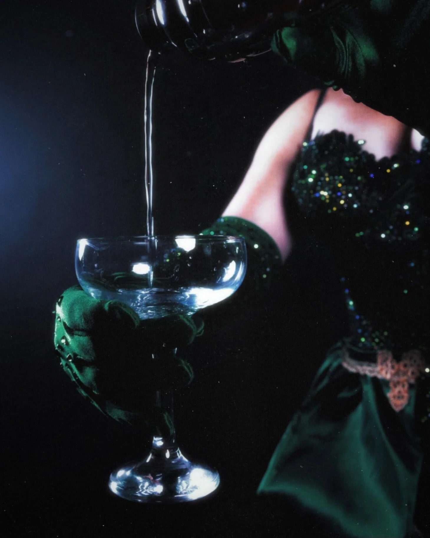 A person wearing green gloves pouring a clear liquid into a glass dessert cup, with a dark background.