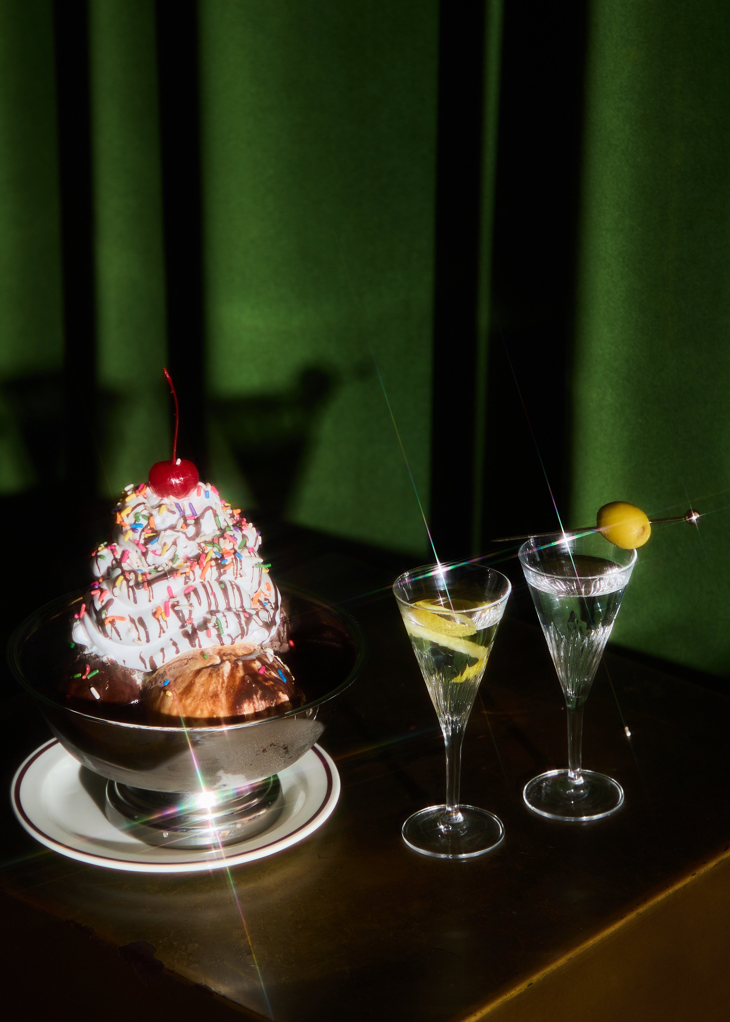 A large dessert with whipped cream, rainbow sprinkles, and a cherry on top in a glass dish, accompanied by two lemon slices in two cocktail glasses, with a green curtain background.