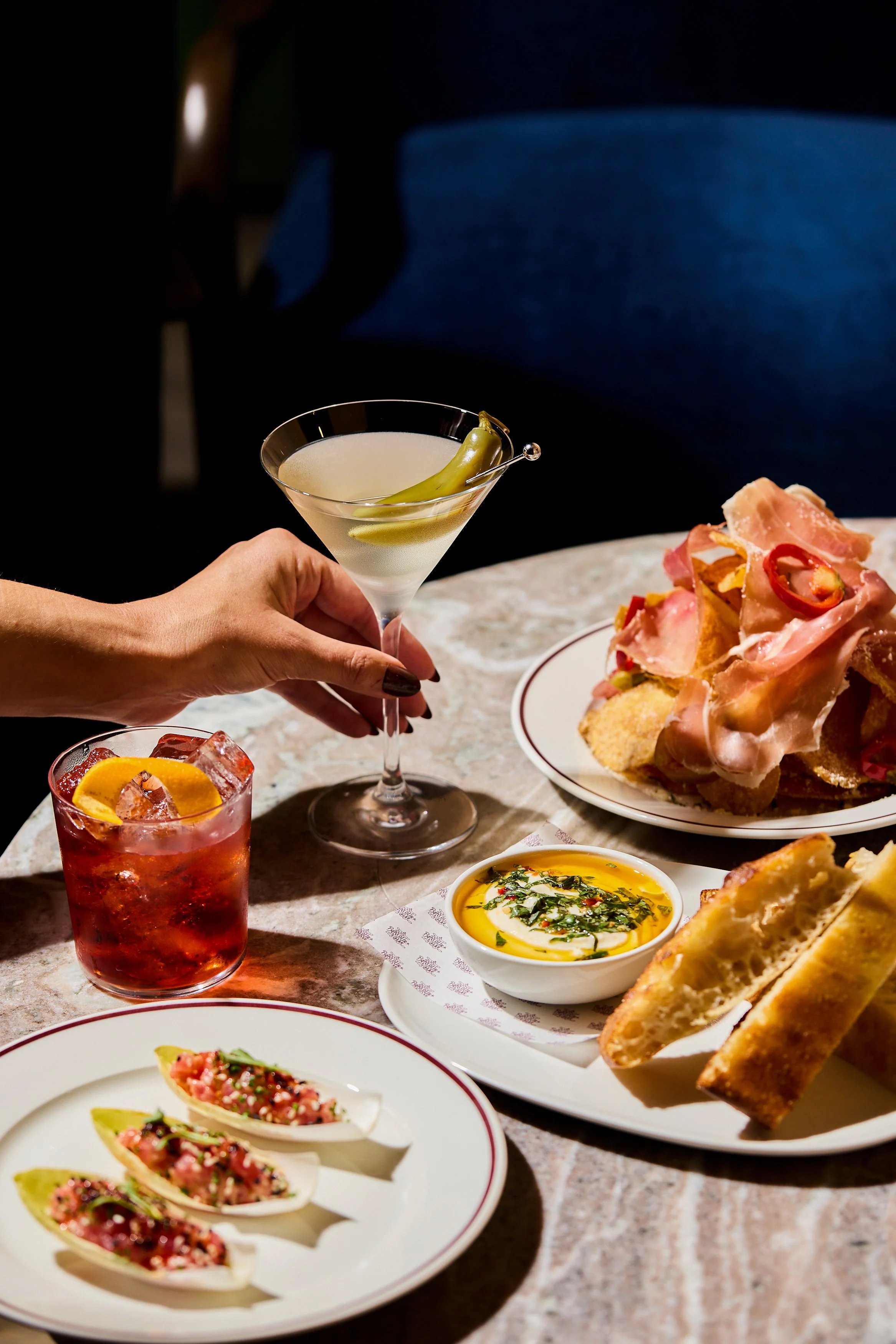 A table with various dishes including a salad on a white plate, a small bowl of olive oil topped with herbs, a sliced baguette, a plate of prosciutto and bread, a glass of pink beverage with an orange wheel and herbs, a martini with a pickle, and ano