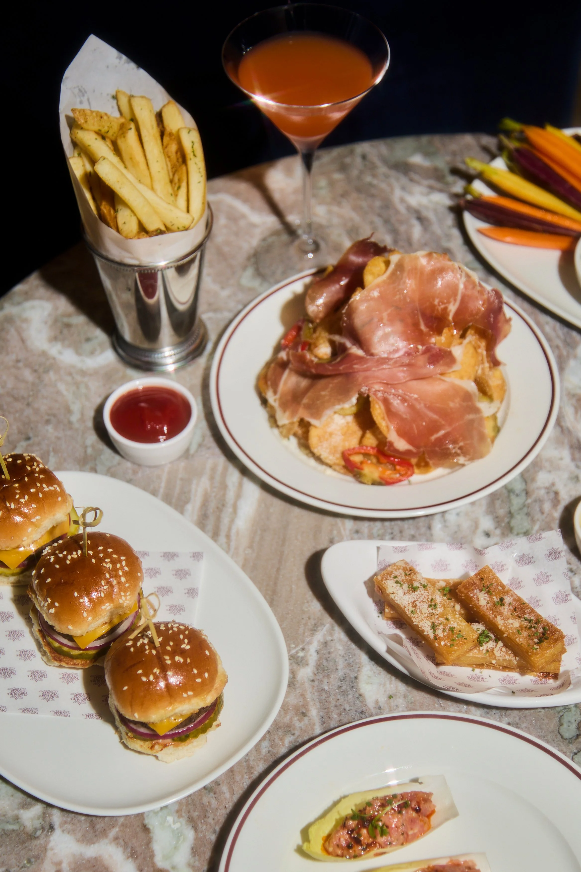 A variety of foods on a table, including French fries in a metal cup, a cocktail in a martini glass, a plate of prosciutto-wrapped appetizers on a bed of chips, a plate with fresh vegetable sticks, a dish with breadsticks topped with herbs, and slide