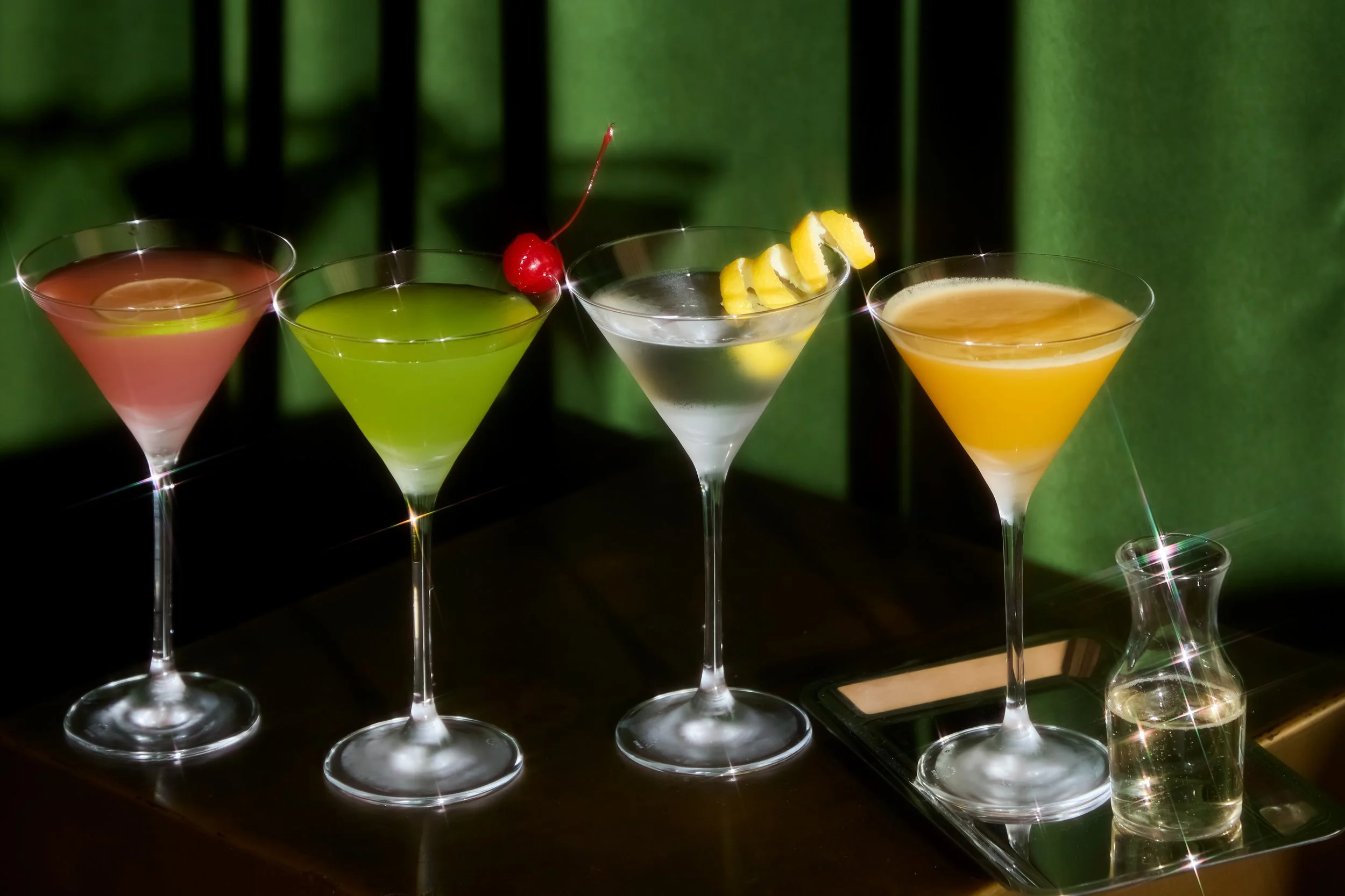 Four colorful cocktails in martini glasses on a dark surface with a green background.