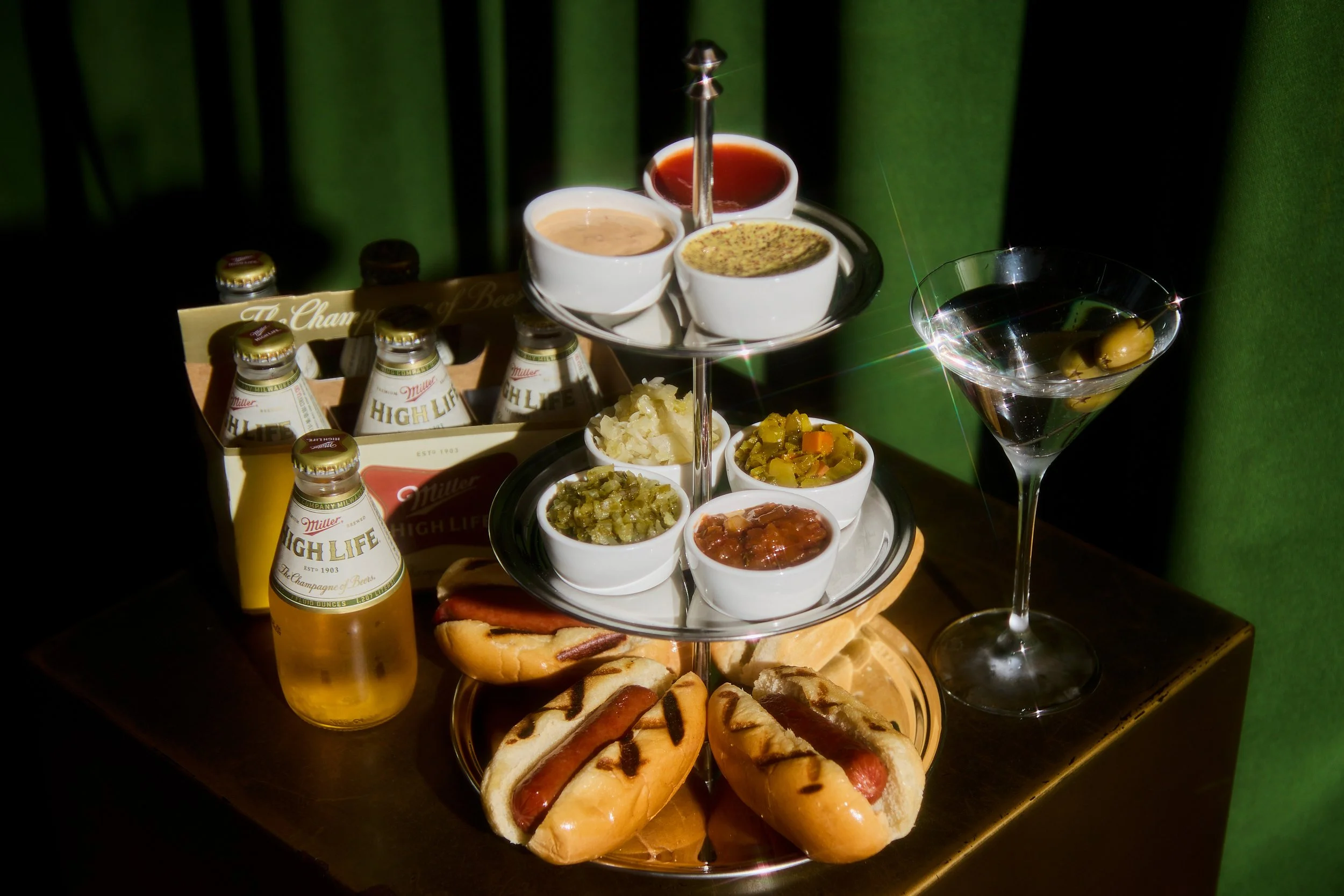A multi-tiered tray with various appetizers, sauces, hot dogs, and soda bottles, set against a green curtain background.