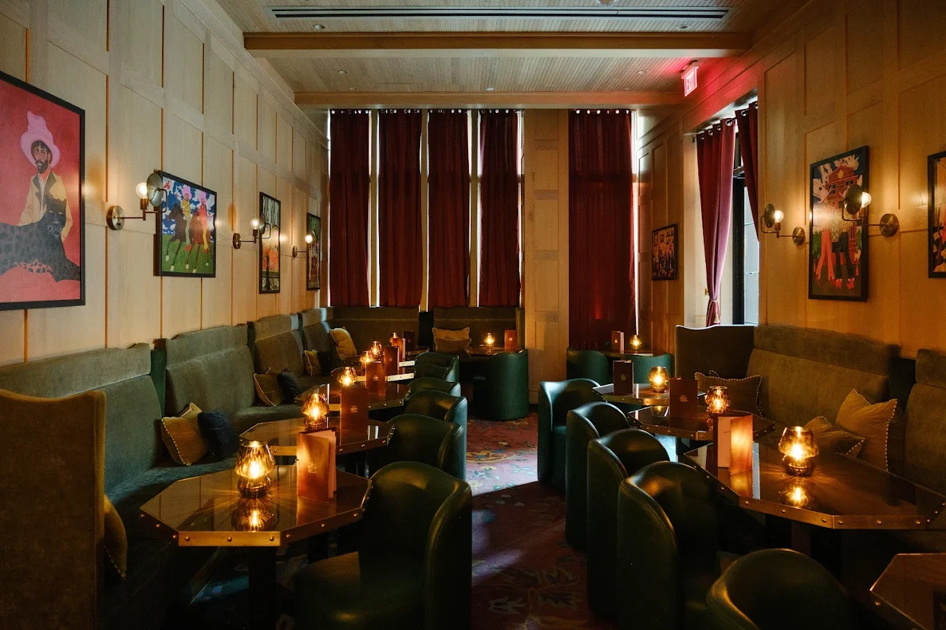 A dimly lit lounge with dark green upholstered seating along the walls, small tables with glowing candles, framed colorful artwork on the wooden-paneled walls, and tall red curtains covering windows.