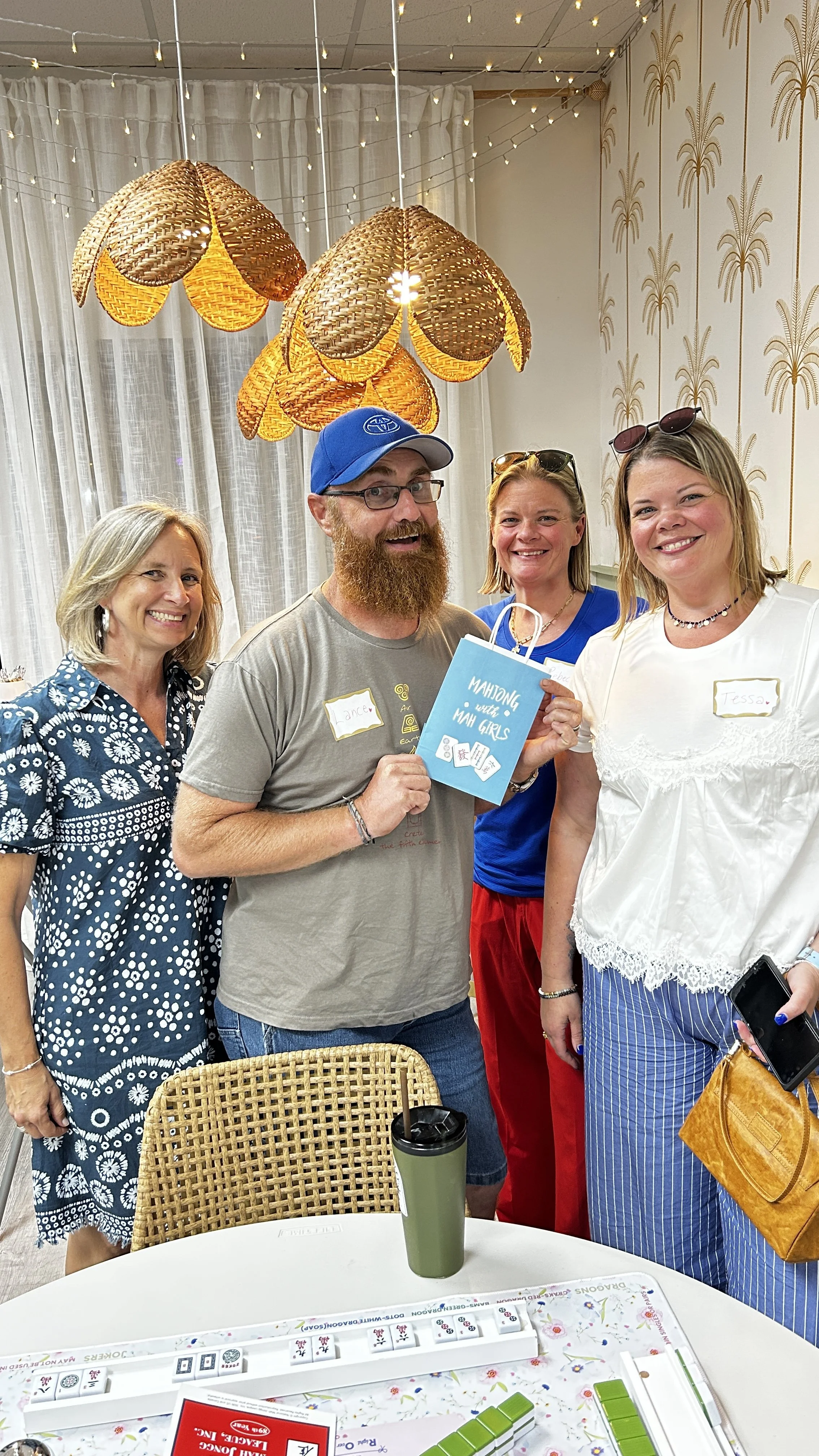 Four people smiling for a photo at a gathering, standing around a table with mahjong game pieces. The man in the center holds a blue bag that says 'Mahjong with Ma Girls.' The room has decorative lighting and wall paper with a palm tree pattern.