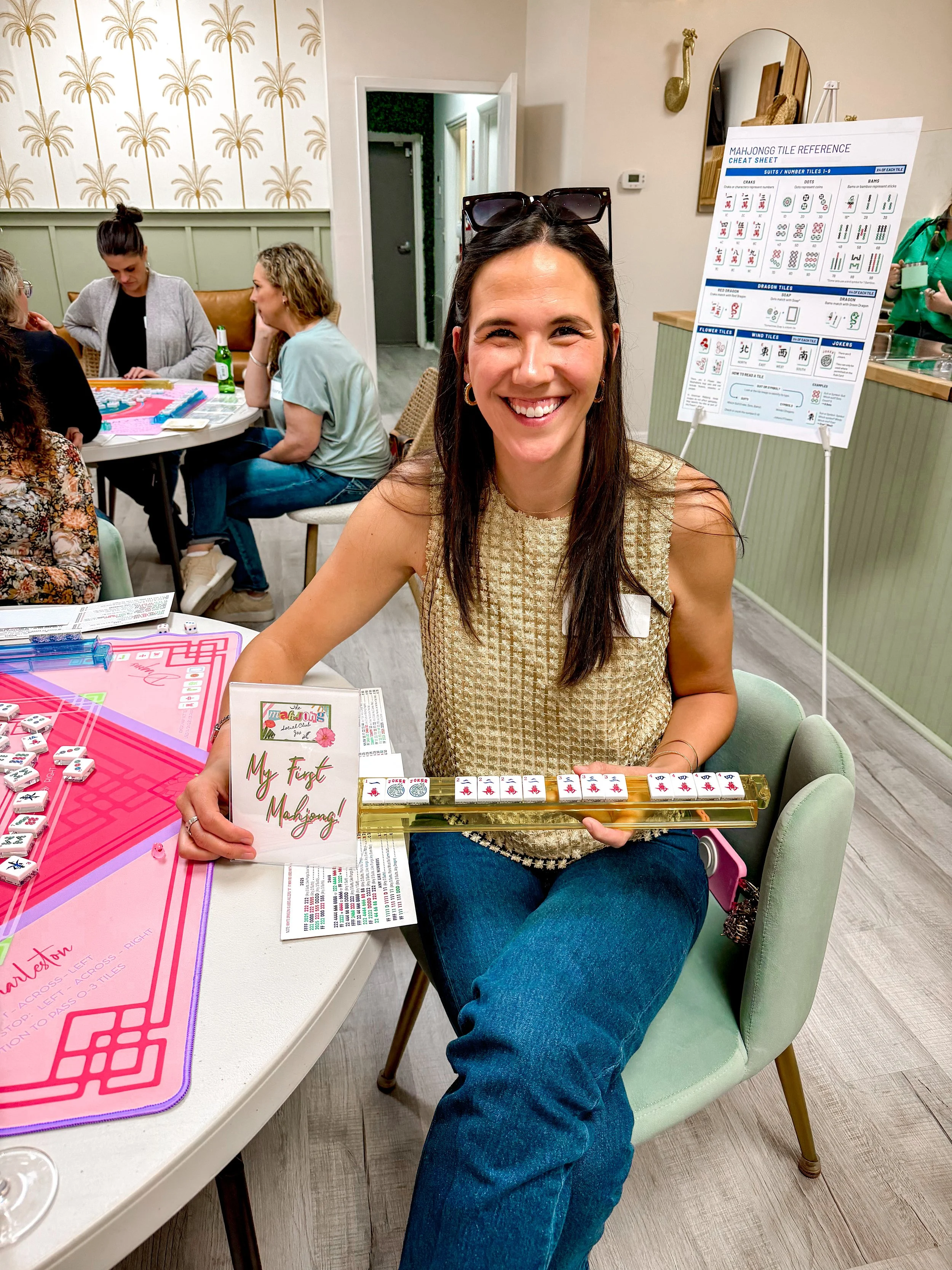 A woman smiling and holding a Mahjong game in a room with other people playing games at a table. There are Mahjong tiles, a sign that says 'My First Mahjong', and a Mahjong reference sheet on an easel in the background.