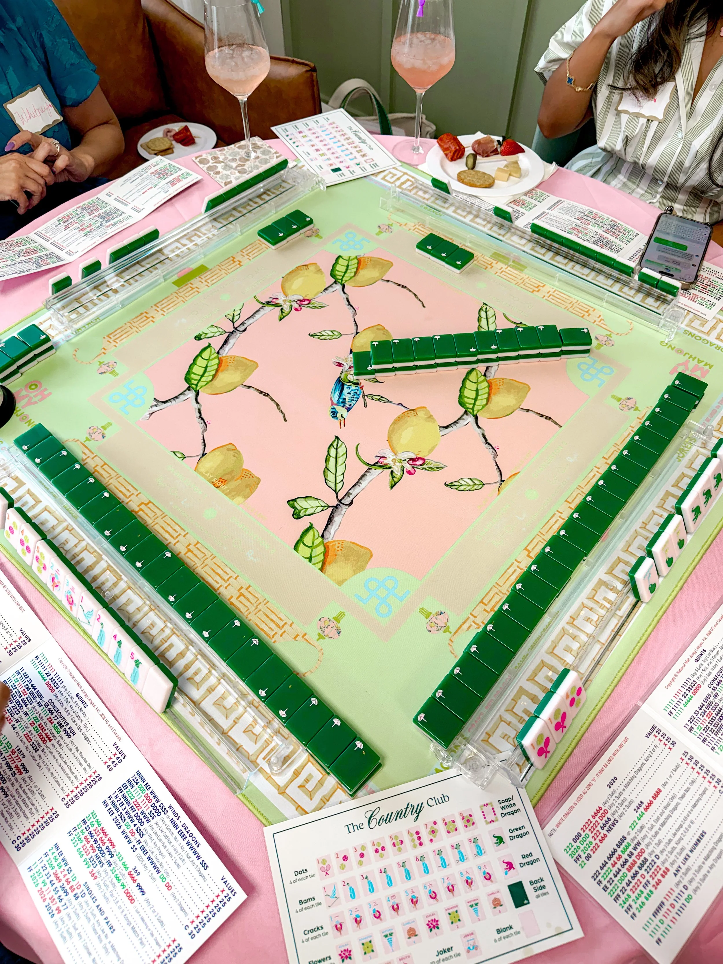 A group of people playing a game of Mahjong around a pink table, with Mahjong tiles on the table and game instructions. Two glasses of pink drinks and plates of snacks are also on the table.