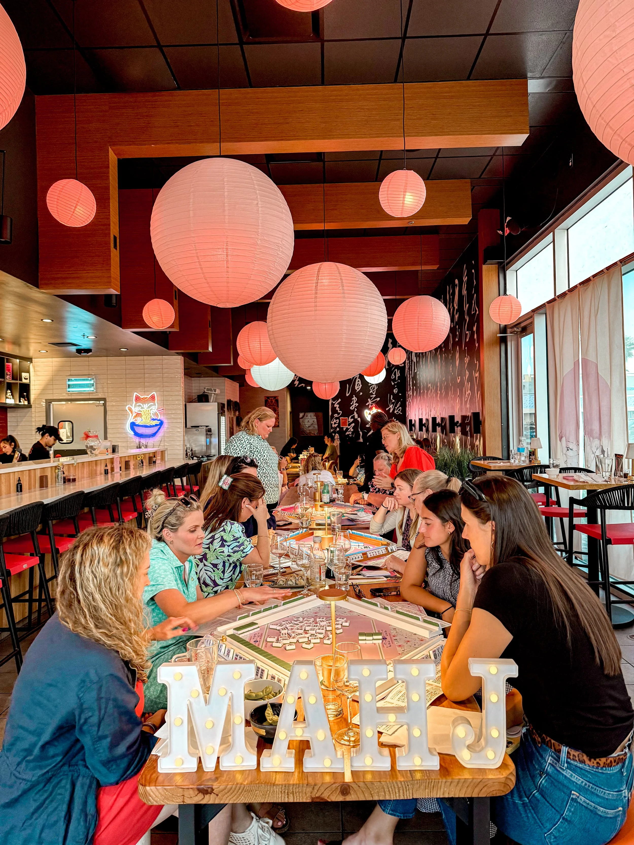 People gathered around a long table playing a game called Mahjong in a brightly lit restaurant with pink paper lanterns hanging from the ceiling. Mahjong open play event.