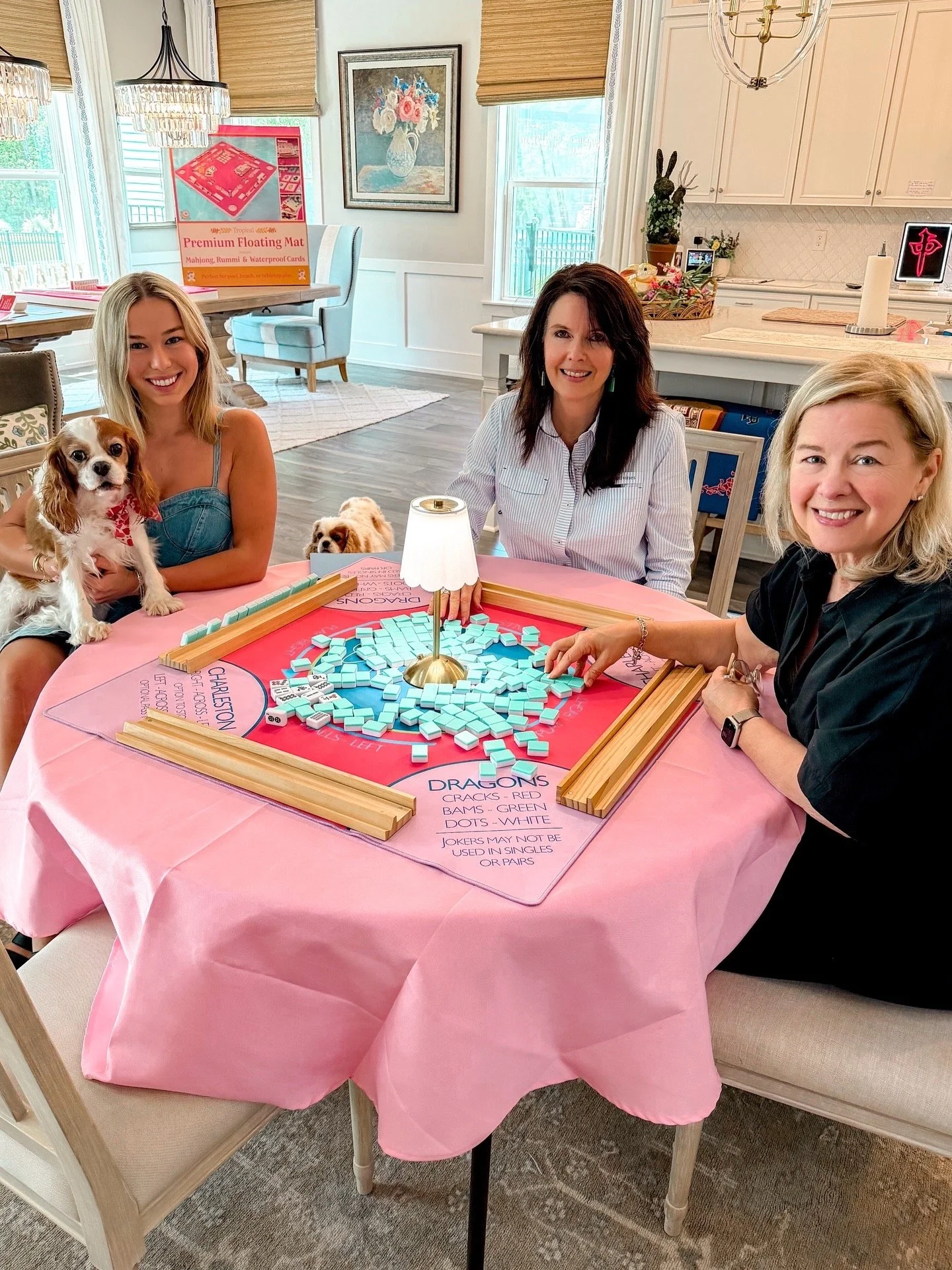 Three women sitting at a table playing dominoes, with two dogs on the woman's lap; the table is covered with a pink tablecloth and a game board for dominoes. The room has large windows, artwork, and kitchen cabinets in the background.