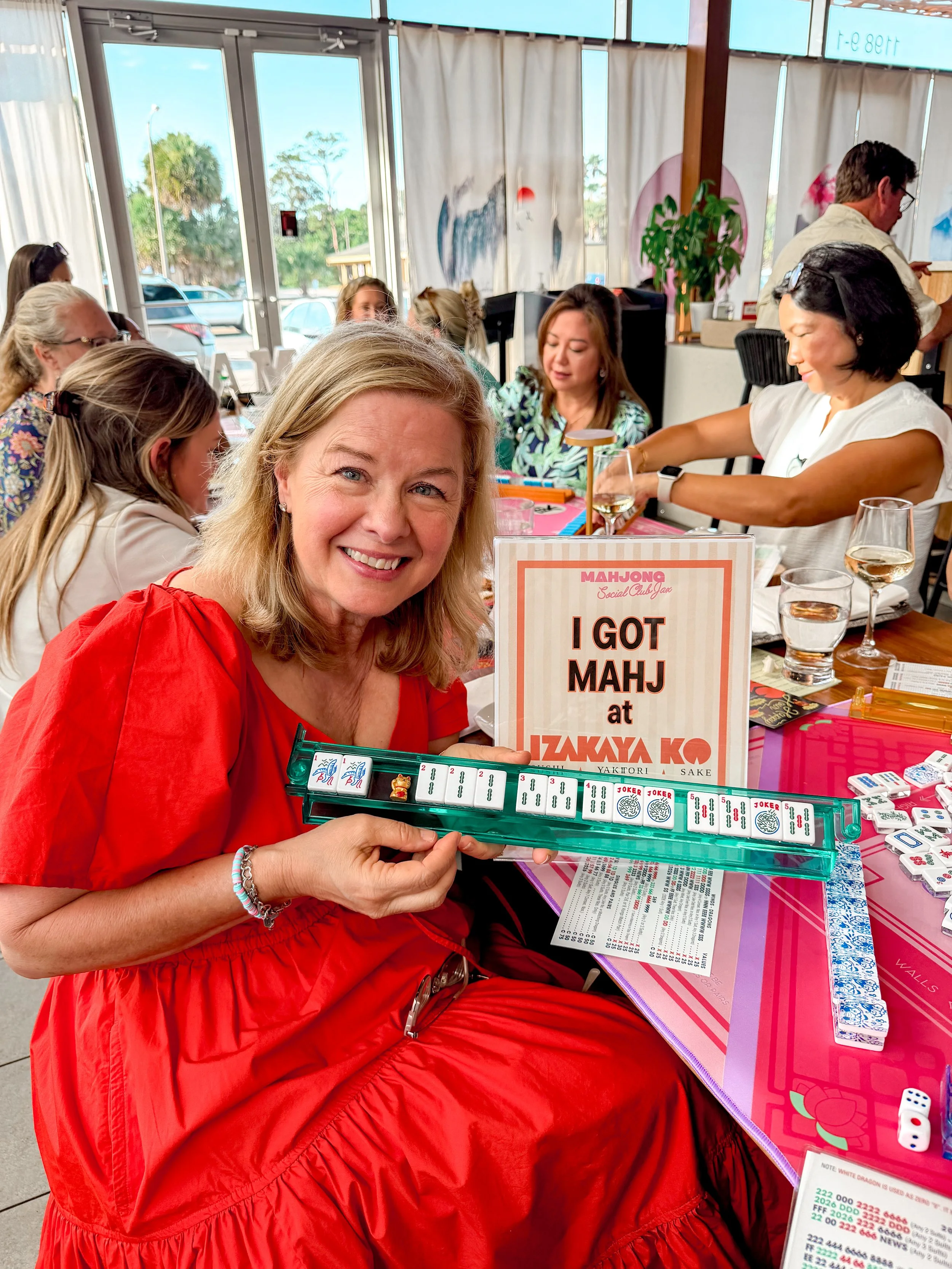 A woman in a red dress smiling and holding a mahjong rack, with mahjong tiles arranged on it, in a restaurant with other people and decorations in the background.