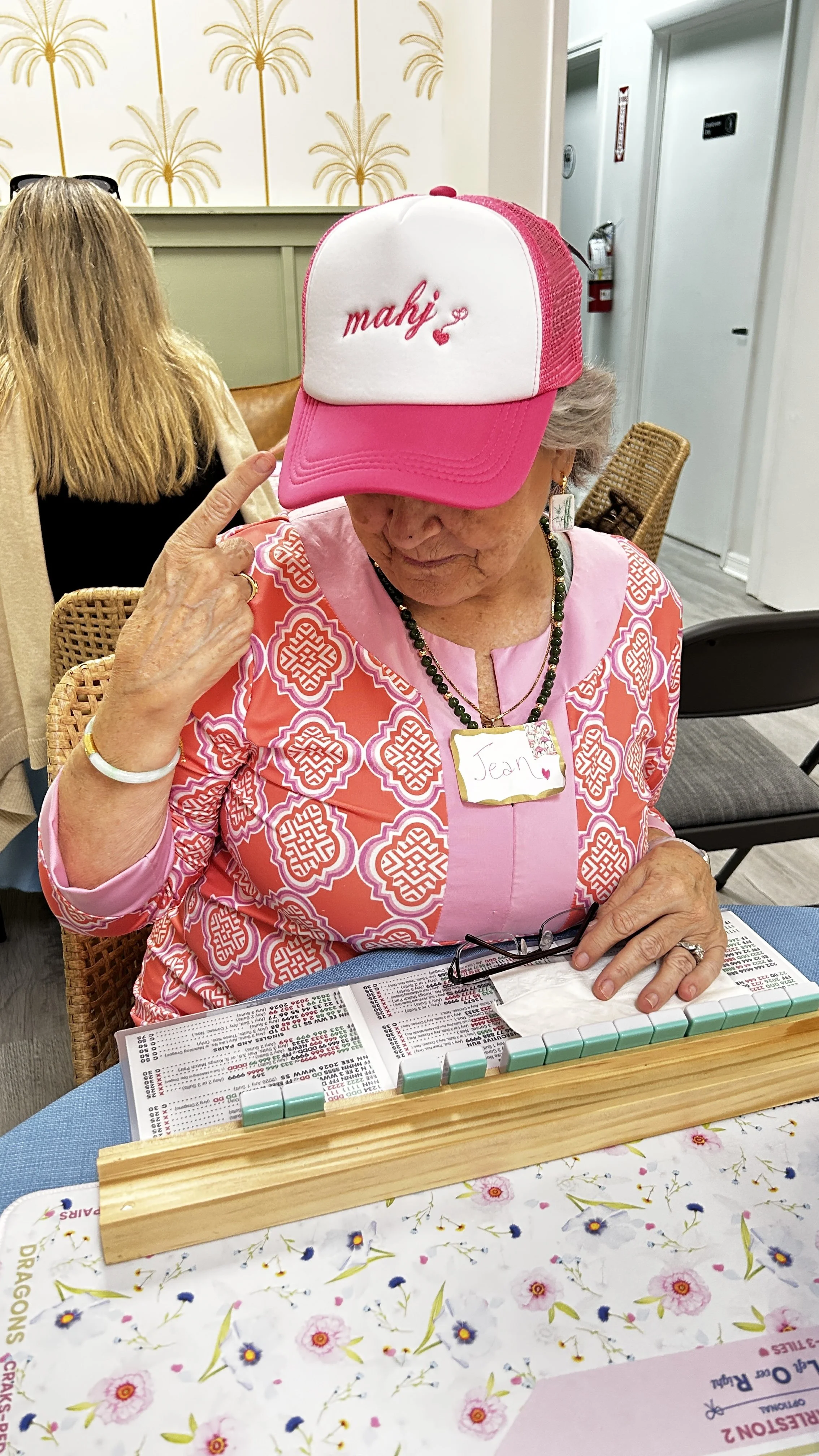 An woman wearing a pink and white patterned top, a pink and white cap with the name "Mahi" embroidered on it, sitting at a table with a MAHJONG game. She has a name tag that says "Jean" and is holding her head with her finger near her temple.