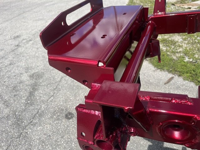 A close-up view of a red-painted metal frame and mounting hardware, likely part of a machine or equipment, outdoors on pavement with some grass in the background.