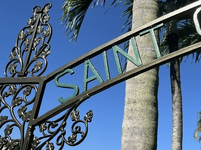 Close-up of an ornate black wrought iron street sign with the word 'SAINT' in colorful letters, against a bright blue sky with palm trees in the background.