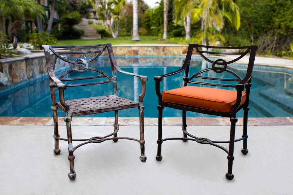 Two metal chairs, one with a wicker seat and the other with an orange cushion, placed by a swimming pool in a backyard with trees and plants.