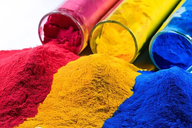 Three glass test tubes lying on their side, each spilling brightly colored powders: red, yellow, and blue.