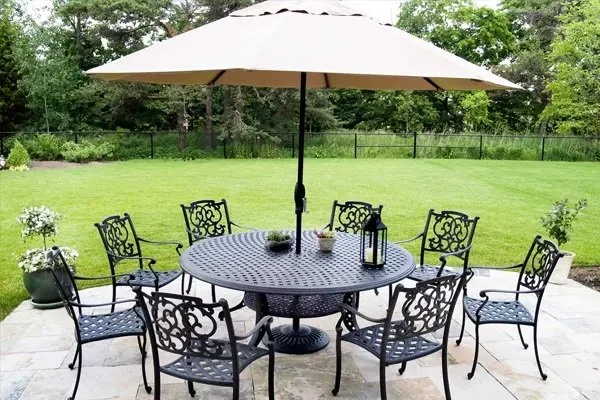Outdoor patio with a round metal table, eight matching chairs, a large beige patio umbrella, and decorative items on the table, set on a tiled surface with a grassy yard and trees in the background.