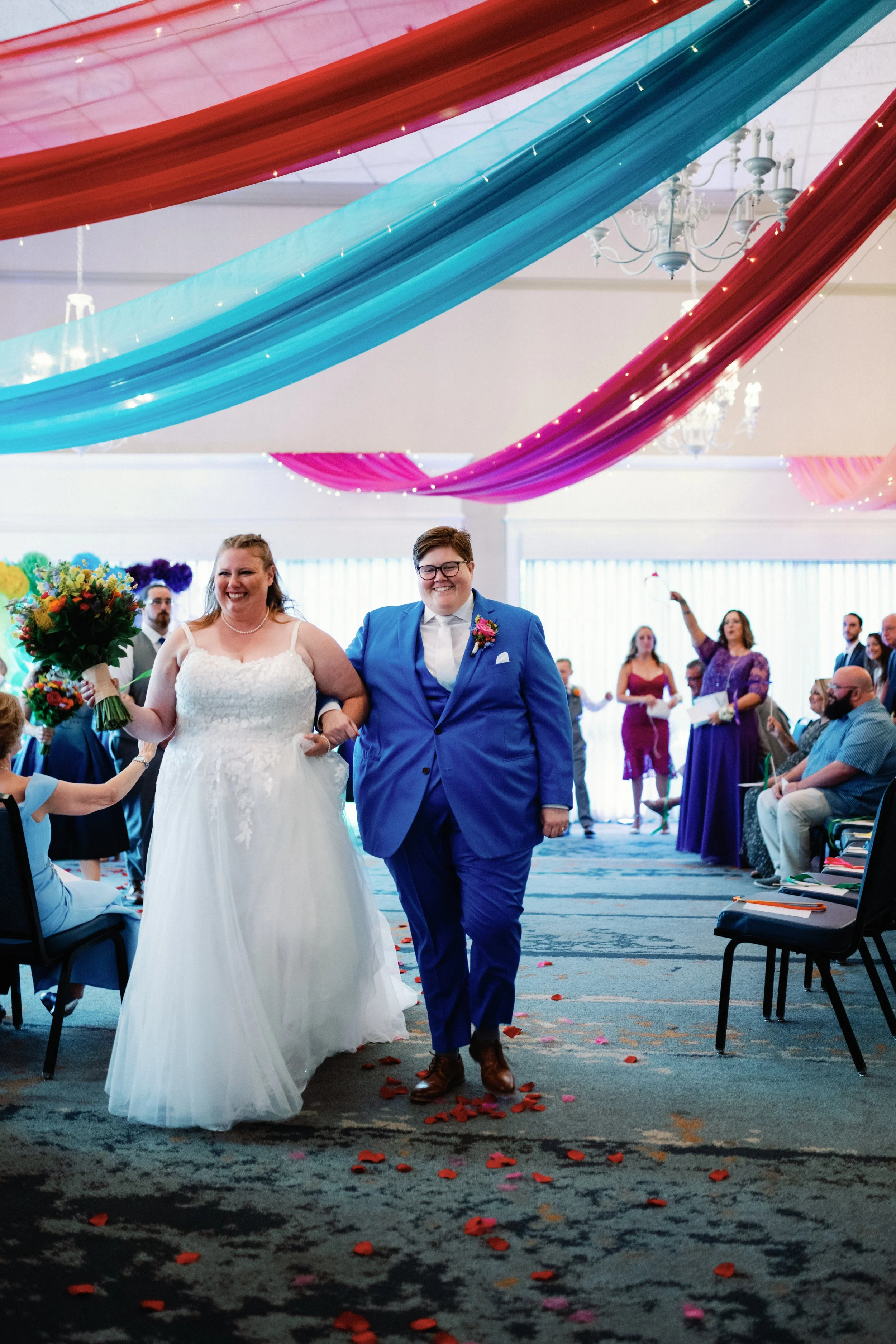 A bride and groom walking down the aisle at their wedding ceremony, surrounded by wedding guests. The bride is wearing a white wedding dress and holding a bouquet, while the groom is dressed in a blue suit. Colorful drapes and flower petals decorate the room.