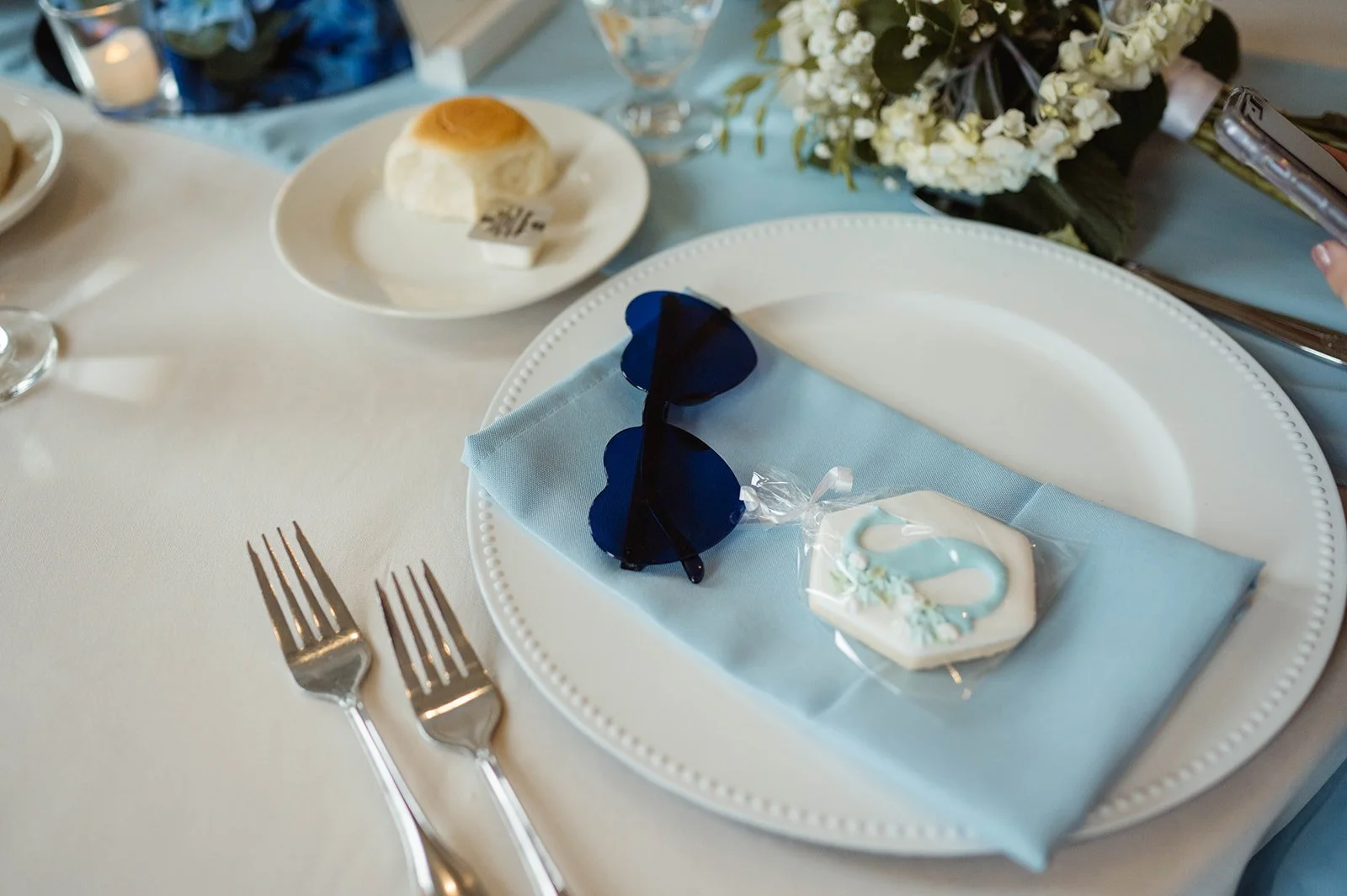 A table setting at a celebration with a white plate, light blue napkin, blue sunglasses, decorated cookie, and small wrapped item, with a floral centerpiece in the background.