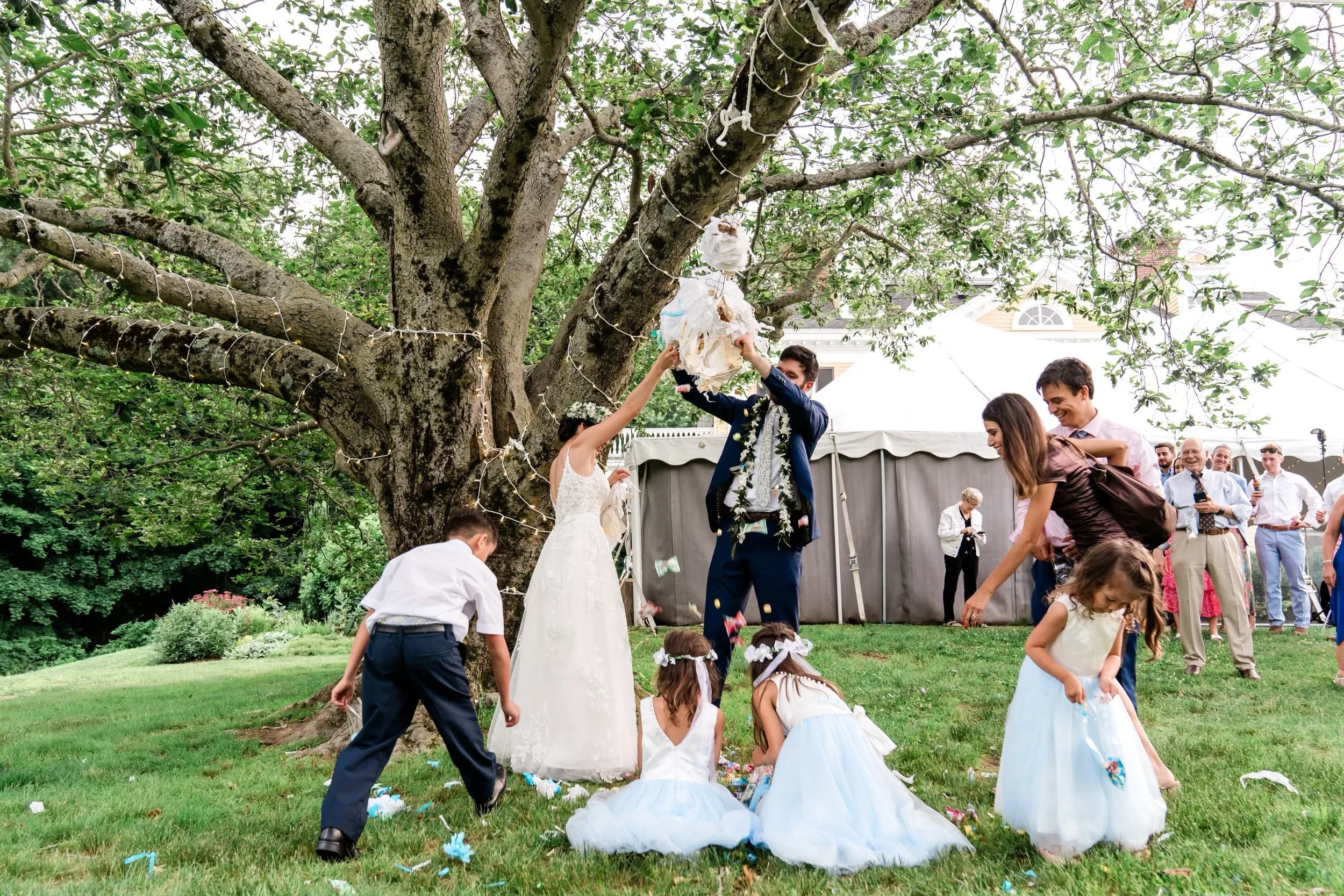 Wedding celebration outdoors under a large tree with string lights, children in white dresses and boys in dark pants, and adults watching. One man and woman are lifting a white object, possibly a piñata, while the children gather around on the grass.