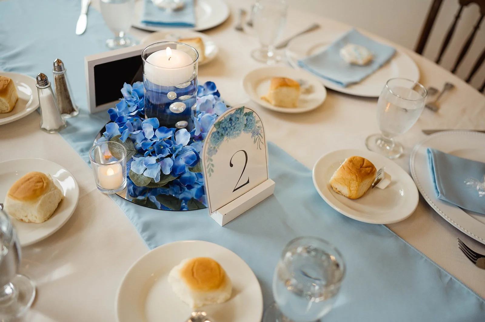 A formal dining table decorated with a centerpiece of a blue candle, artificial blue hydrangea flowers, and small candles. The table has white plates with bread rolls, clear glasses of water, blue napkins, and silverware. There is a table number, '2'