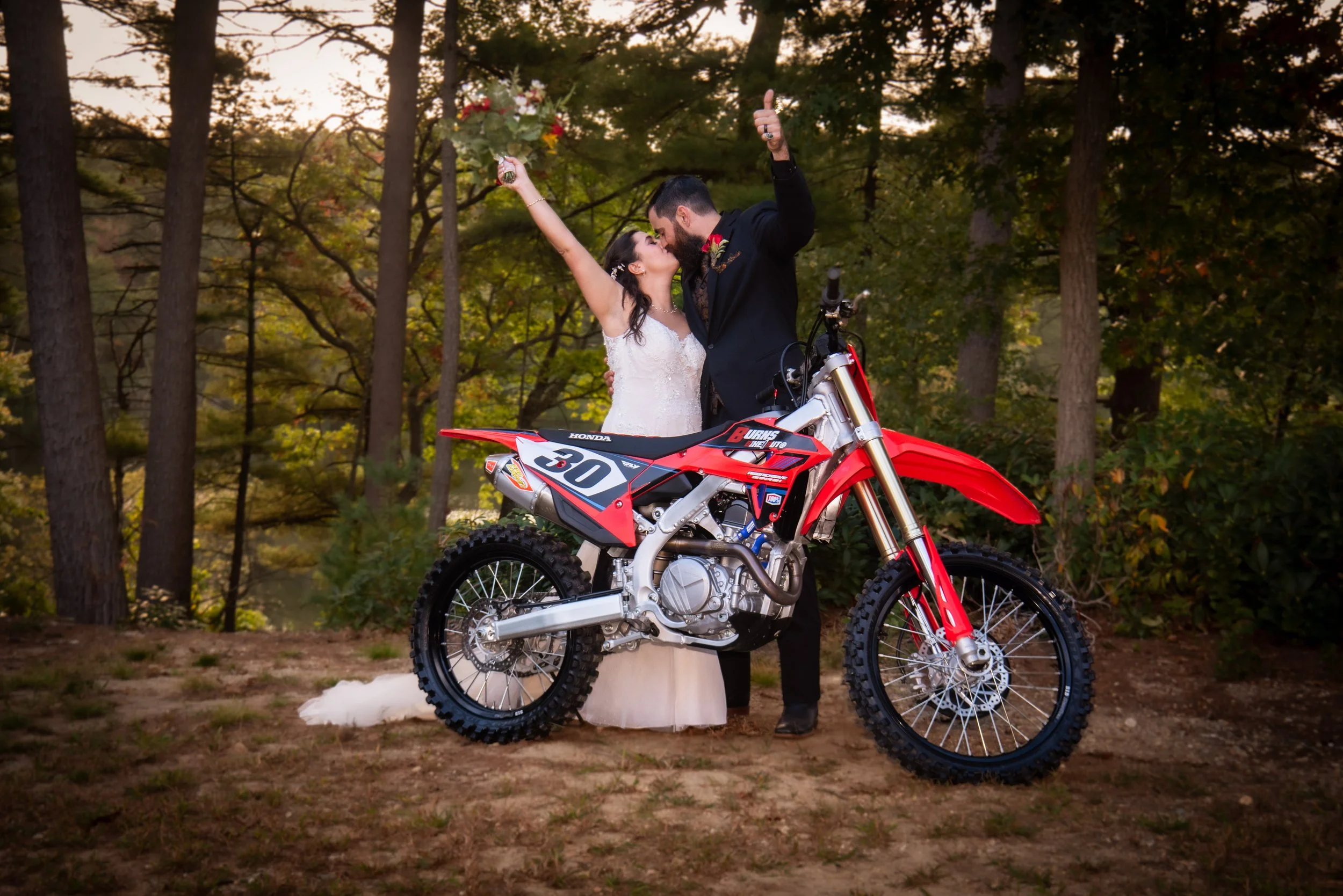 A bride and groom sharing a kiss in a wooded outdoor setting, with the groom standing behind a red and white motocross bike, and the bride holding a bouquet of flowers.