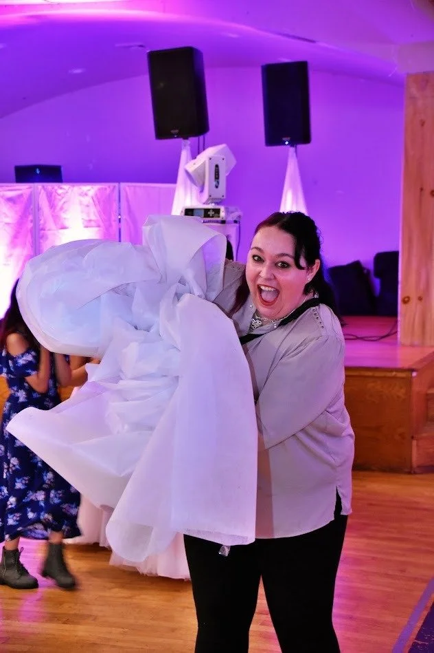 A woman smiling and holding large white fabric at an indoor event with purple lighting, speakers, and a wooden stage in the background.